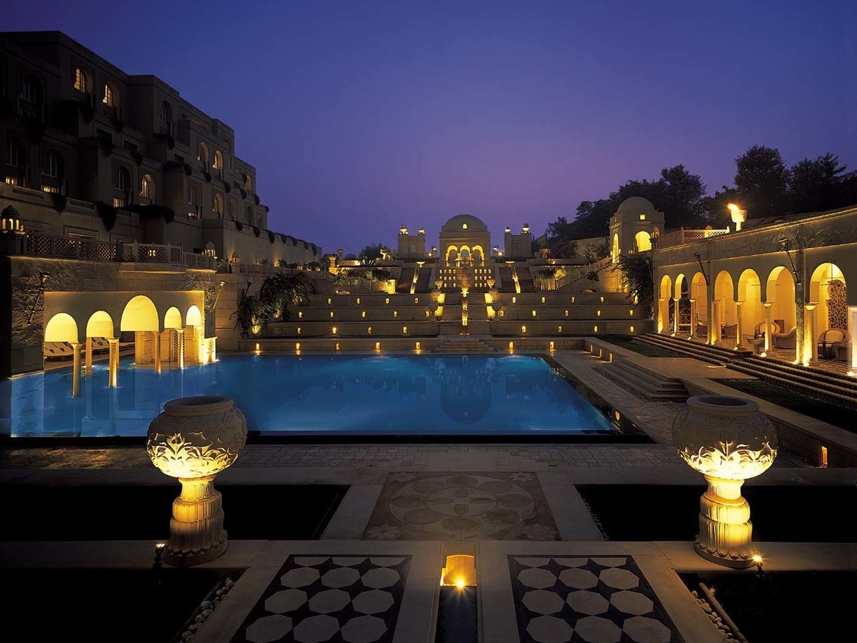 An illuminated swimming pool and tiered stone architecture at night under a deep purple sky.