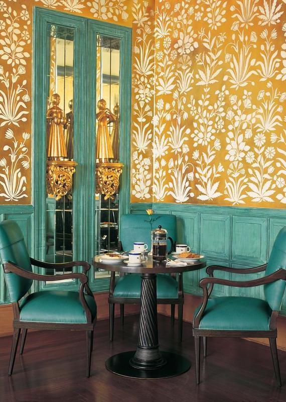 A small dining table with teal chairs against gold floral patterned walls and mirrored panels.