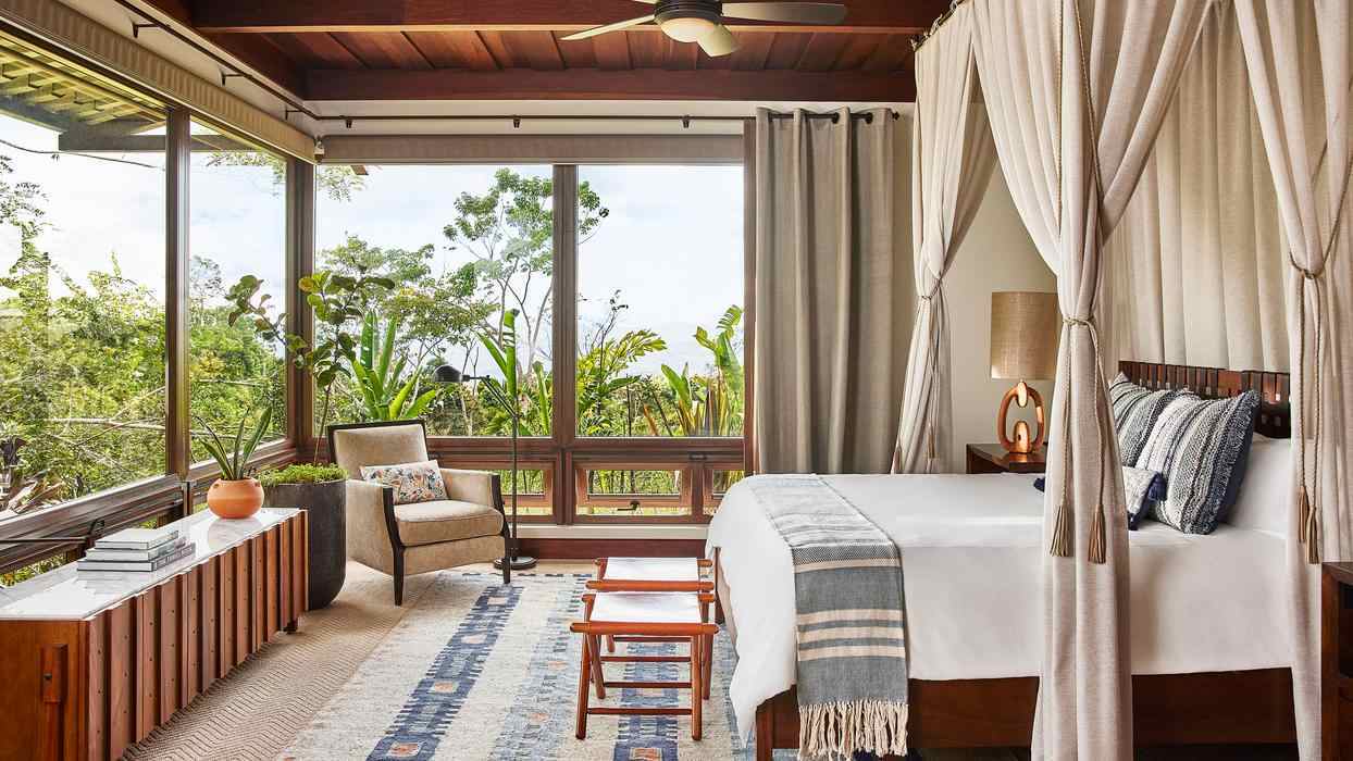 A luxury bedroom with a four-poster canopy bed and floor-to-ceiling windows showing a lush tropical forest.