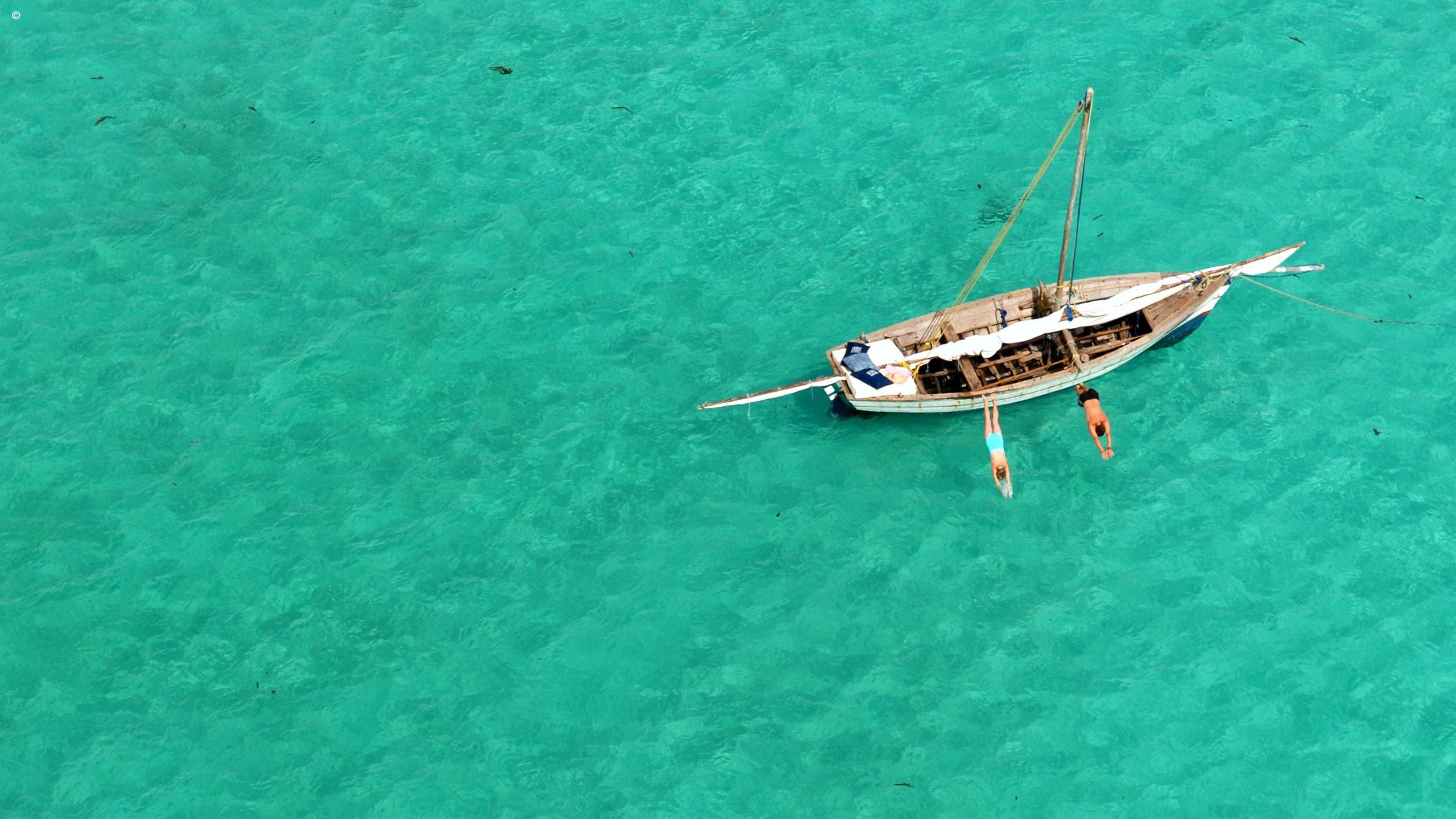 Aerial view of a small boat anchored in crystal-clear turquoise water, with two people diving off the side.