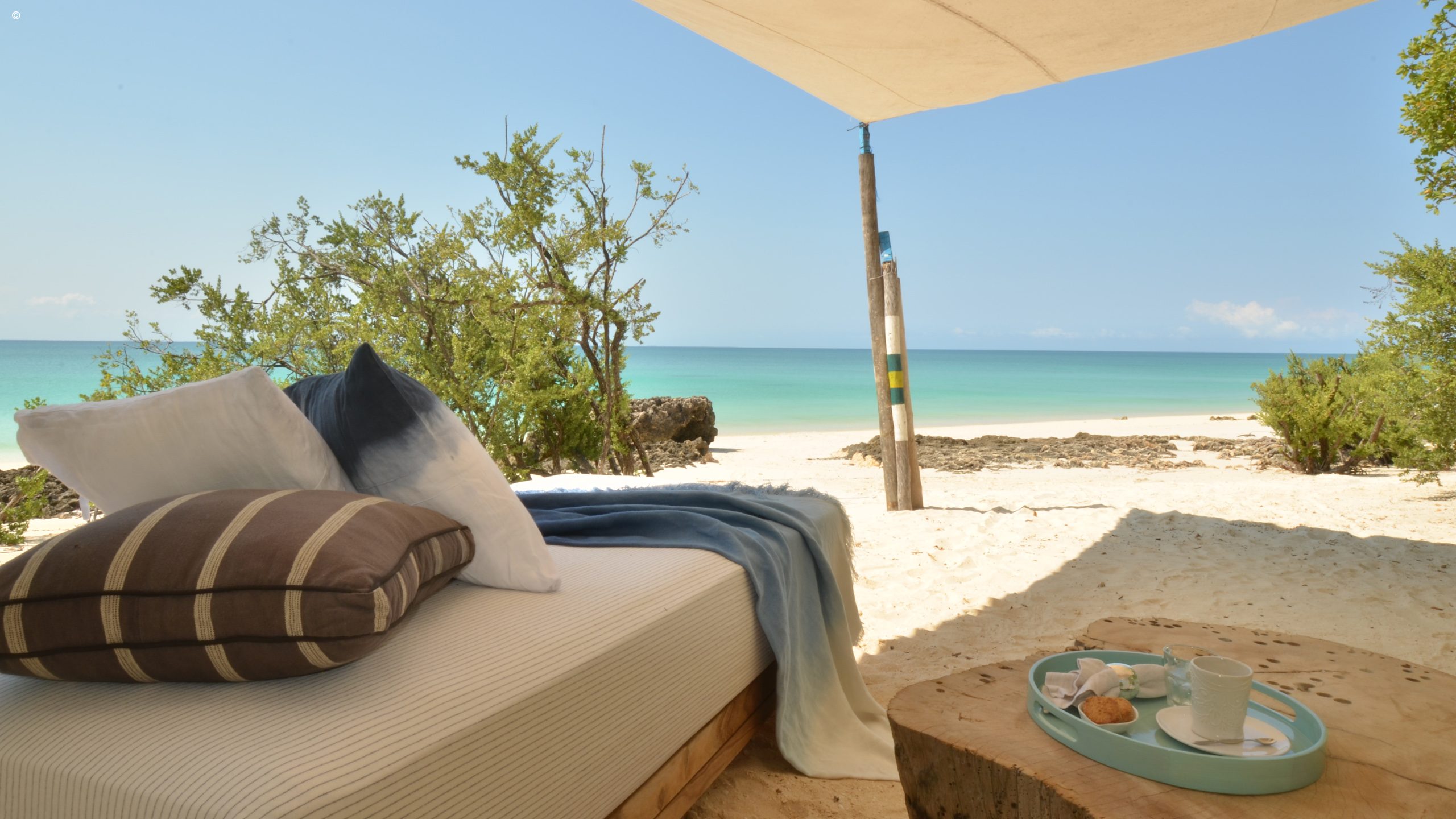 A serene beach view from a shaded lounge area with plush pillows, a cozy blanket, and a tray of refreshments.