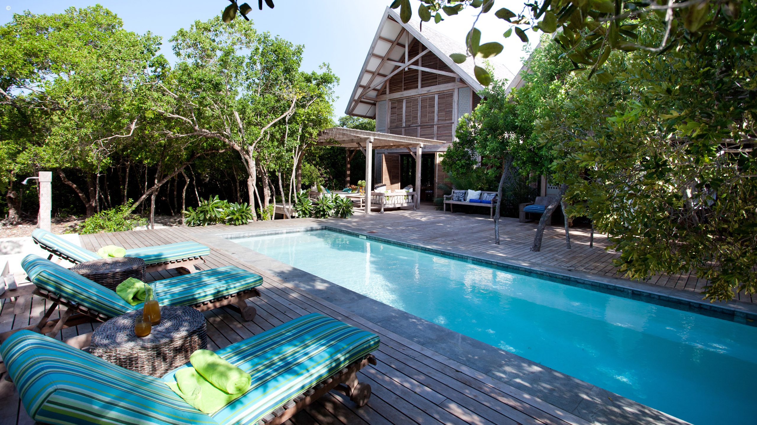 A serene outdoor pool area with striped lounge chairs, a wooden deck, and a stylish house surrounded by lush green trees.
