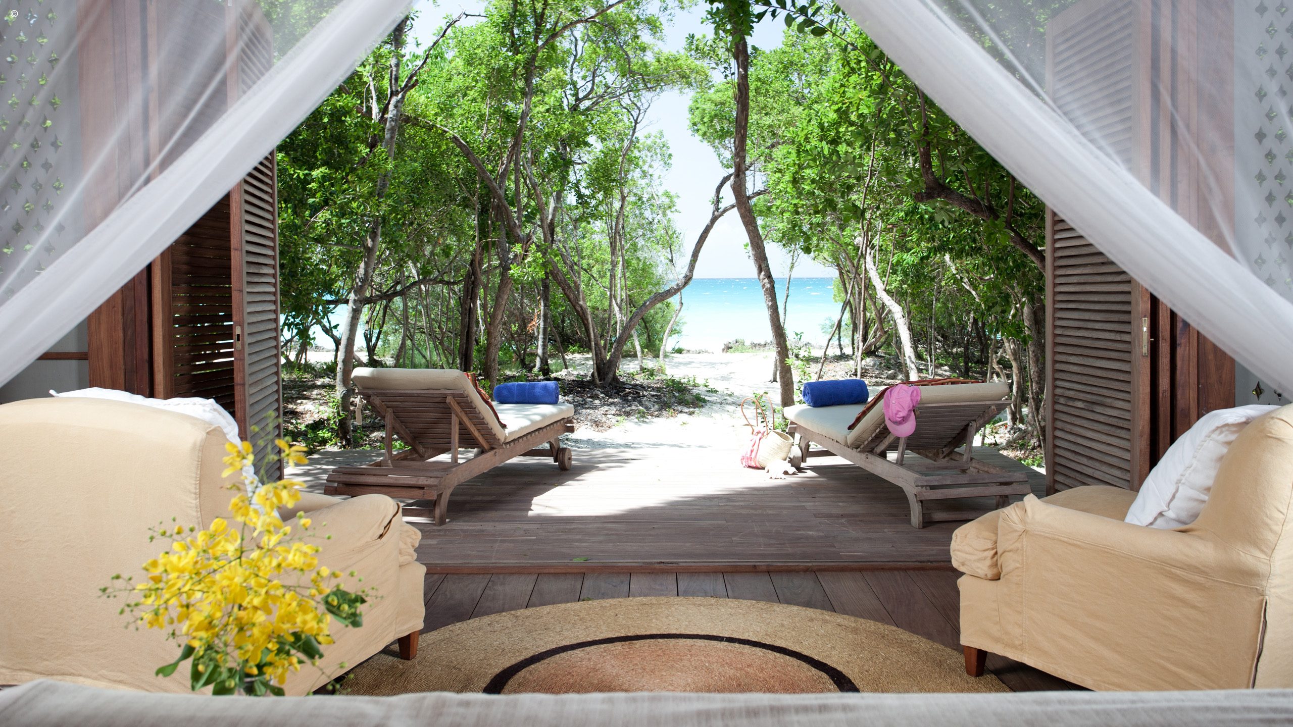 A cozy outdoor view from a luxurious room, featuring lounge chairs, lush greenery, and a glimpse of the beach and ocean beyond.