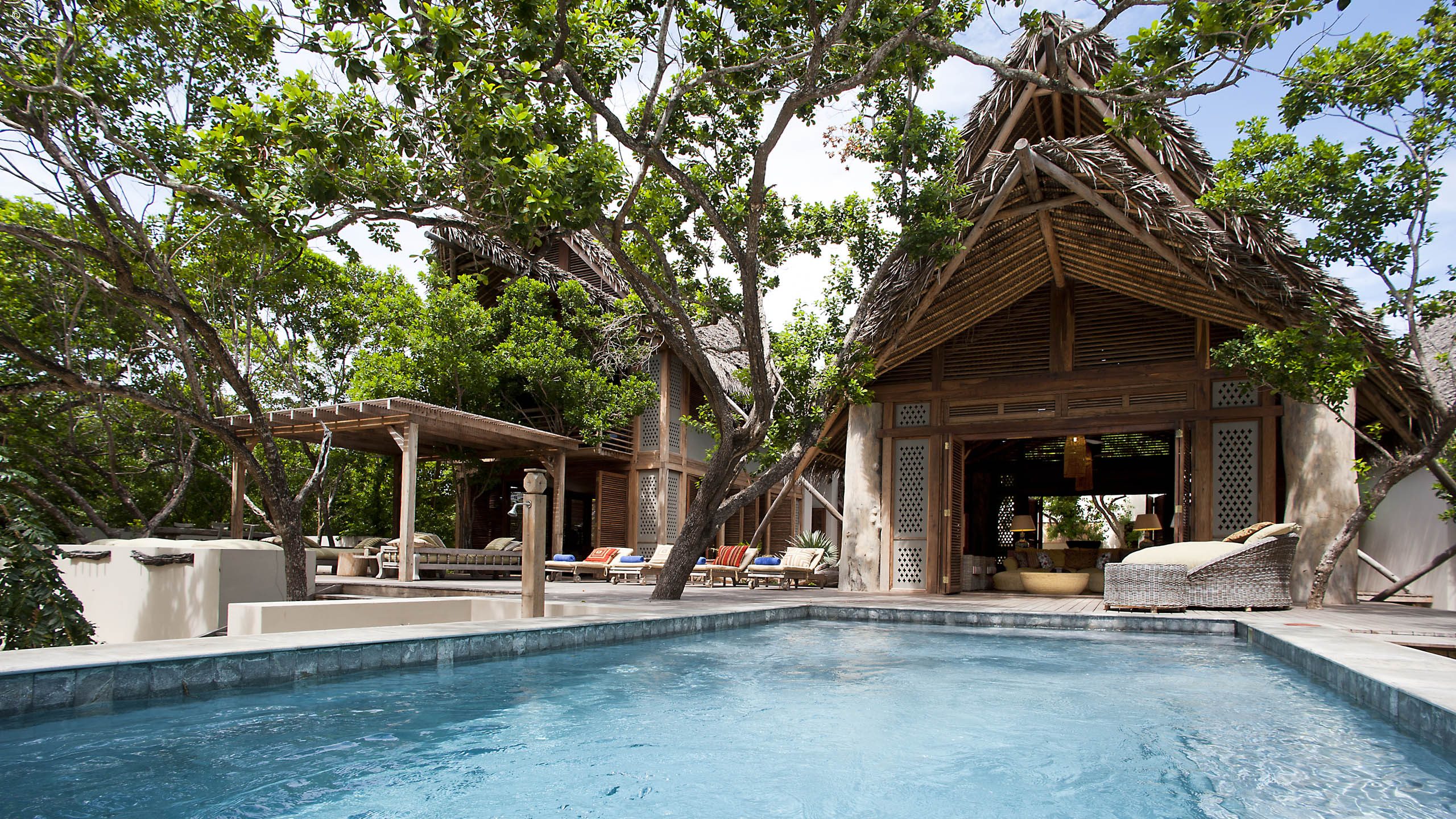 A serene villa with a thatched roof, surrounded by lush greenery and featuring a blue swimming pool, inviting relaxation and tranquility.