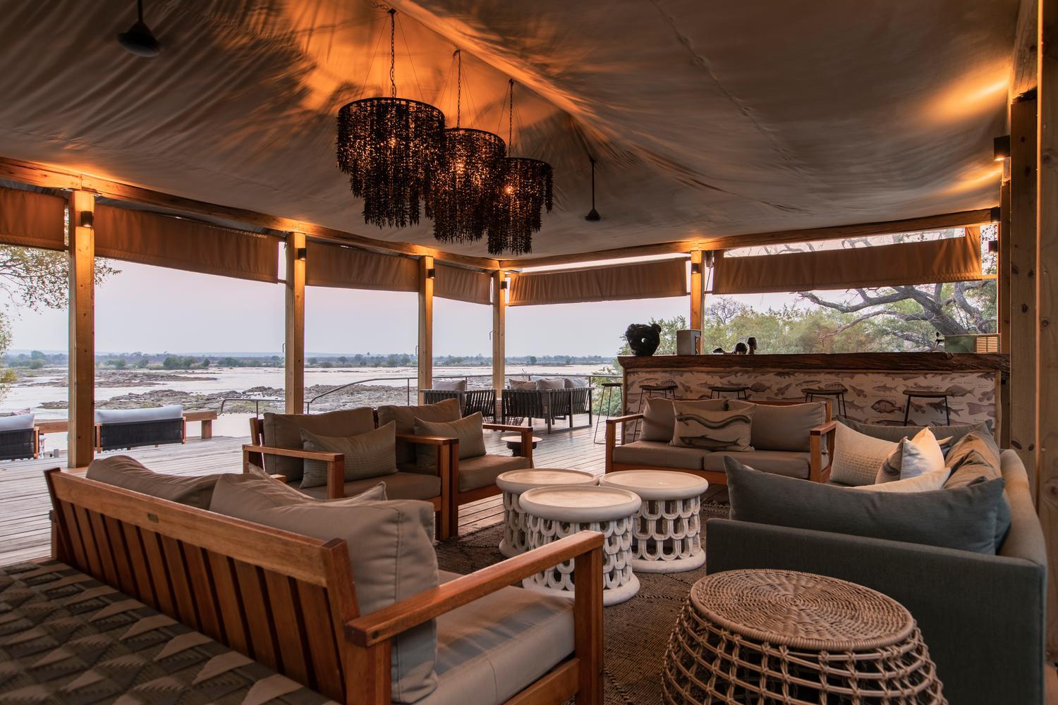 Open-air lounge under a canvas canopy featuring beaded chandeliers, plush wooden seating, a bar, and a view of a wide river.