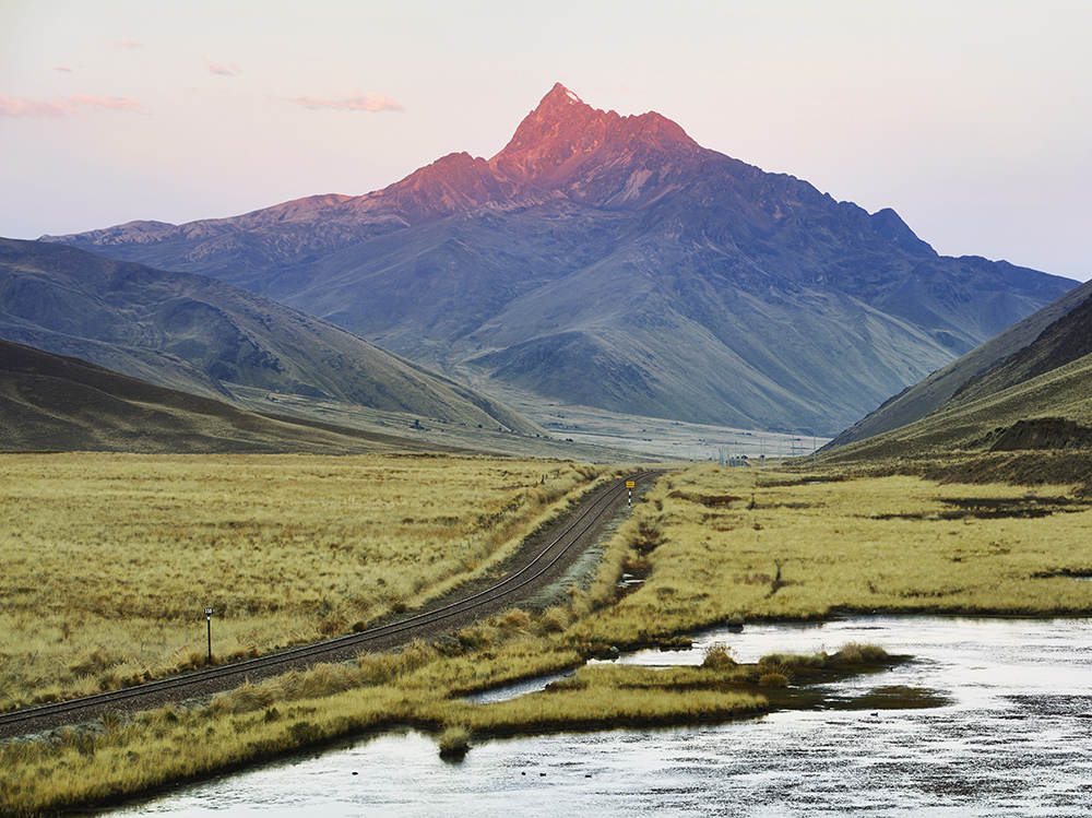 A glowing mountain peak at sunset overlooking railway tracks in a grassy valley with a stream.