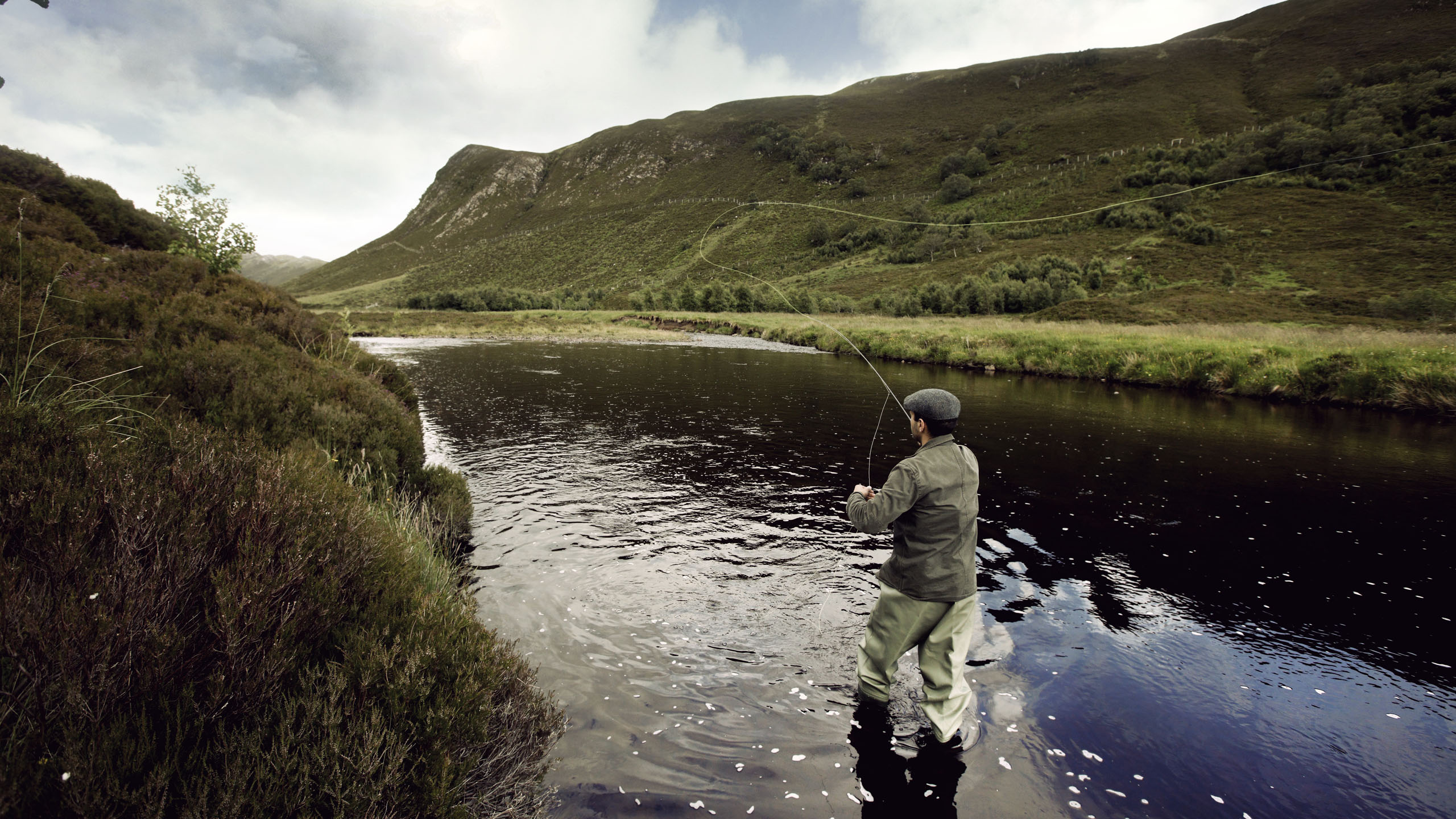A man fly fishing in a river surrounded by rolling green hills under an overcast sky.