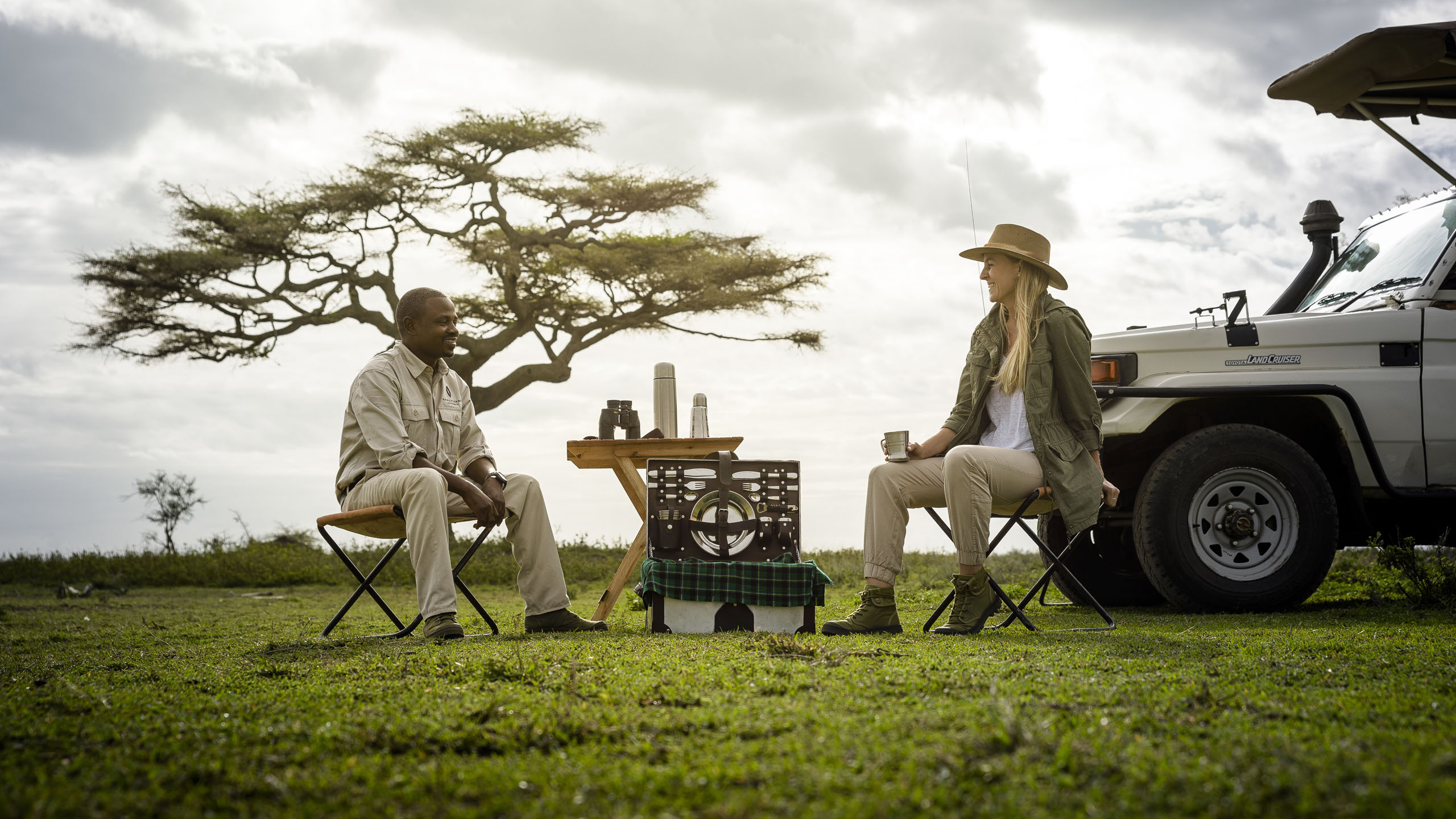 Two people witting around a table drinking coffee by a safari jeep