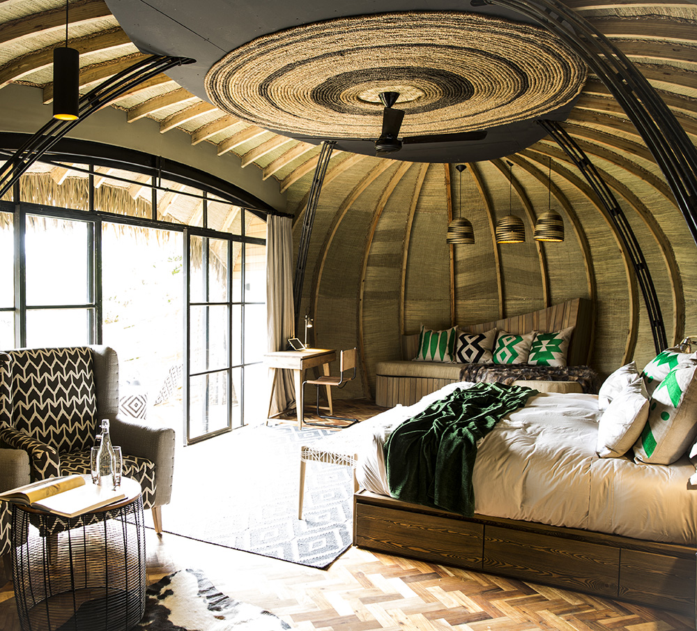 Interior of a luxury lodge with a large bed, woven ceiling decor, and glass doors leading to a balcony.