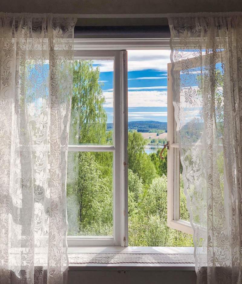 Looking through an open window with white lace curtains at a sunny green forest and river.