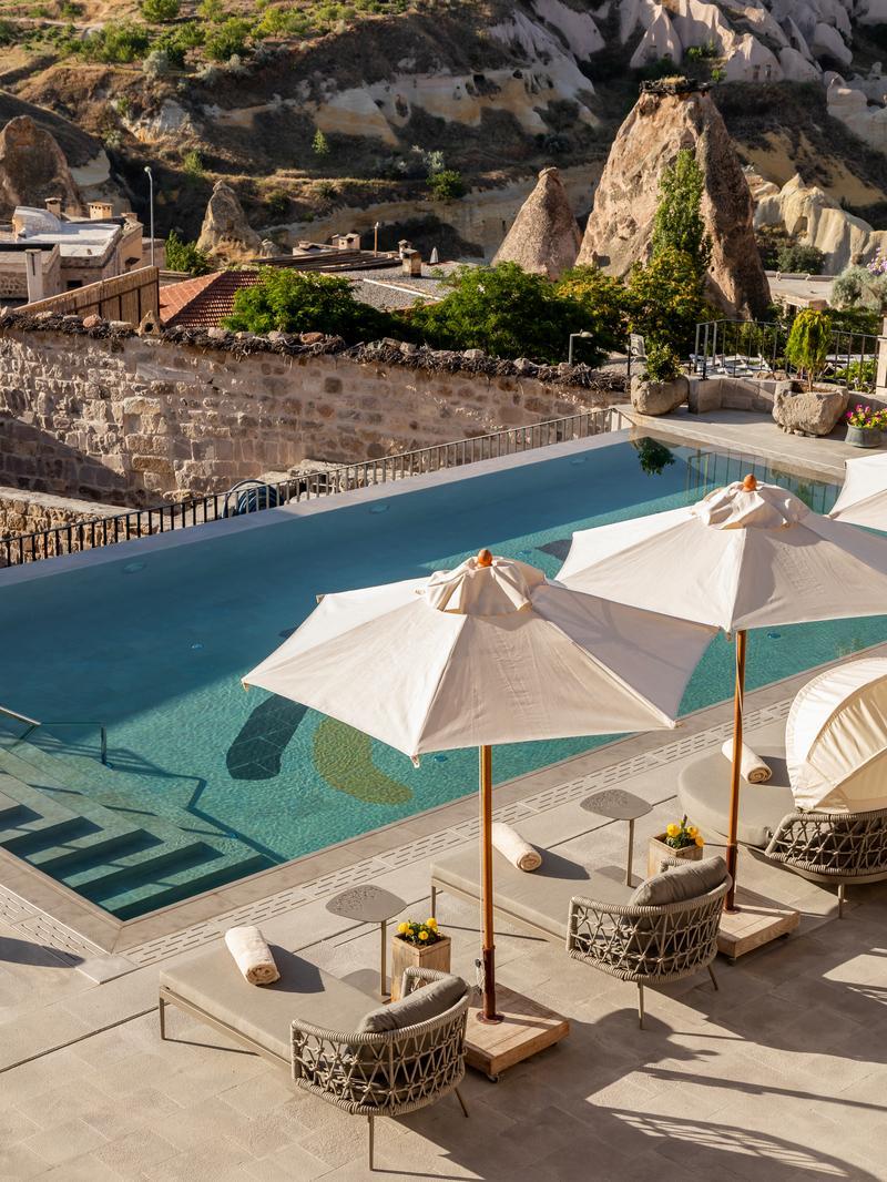 A blue infinity pool with sun loungers and umbrellas overlooking a rugged Turkish landscape.