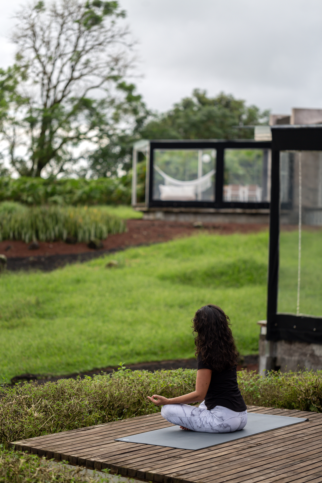 Woman meditating outdoors on a wooden deck with greenery and a modern structure in the background.