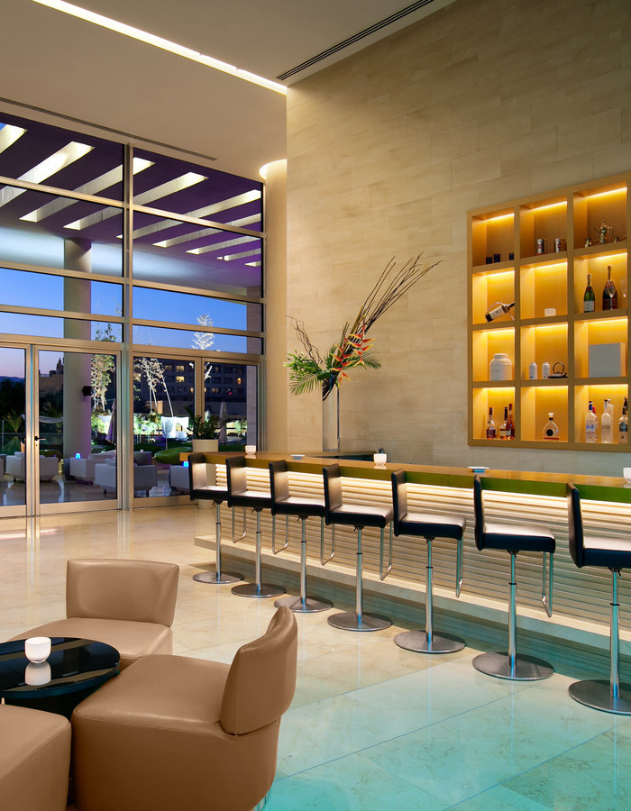 Modern bar area at Kempinski Hotel Aqaba featuring black stools, lit shelves with bottles, and glass walls facing outdoors.