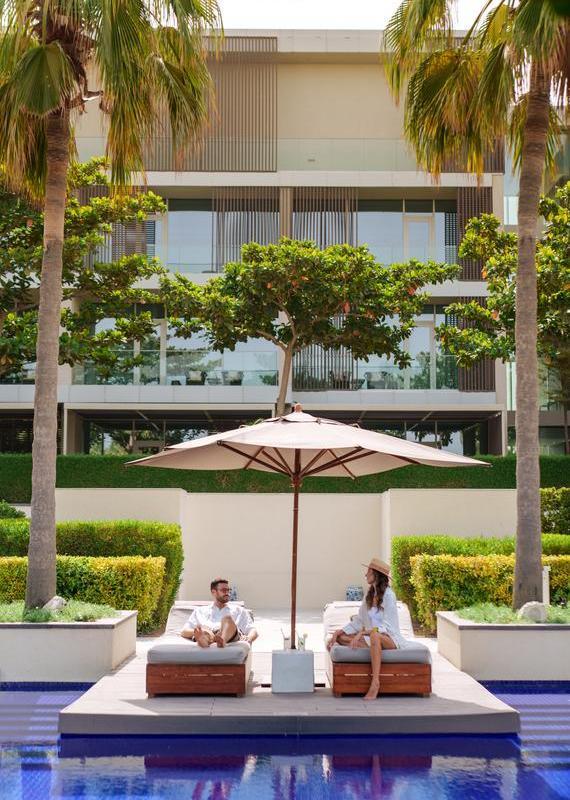 A man and woman sit on lounge chairs under a white umbrella between palm trees by a blue swimming pool.