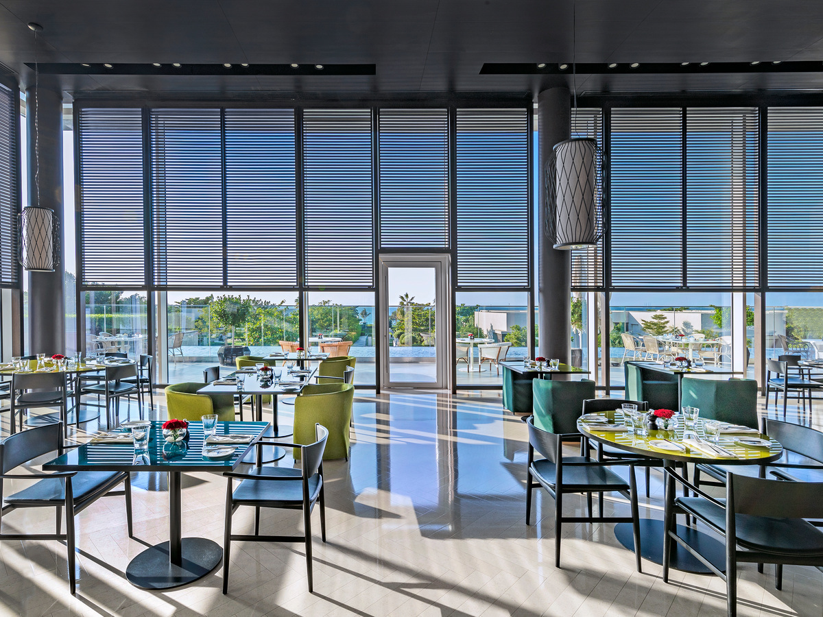 Modern dining room with glass walls, sunlight patterns on the floor, and set tables overlooking the landscape.