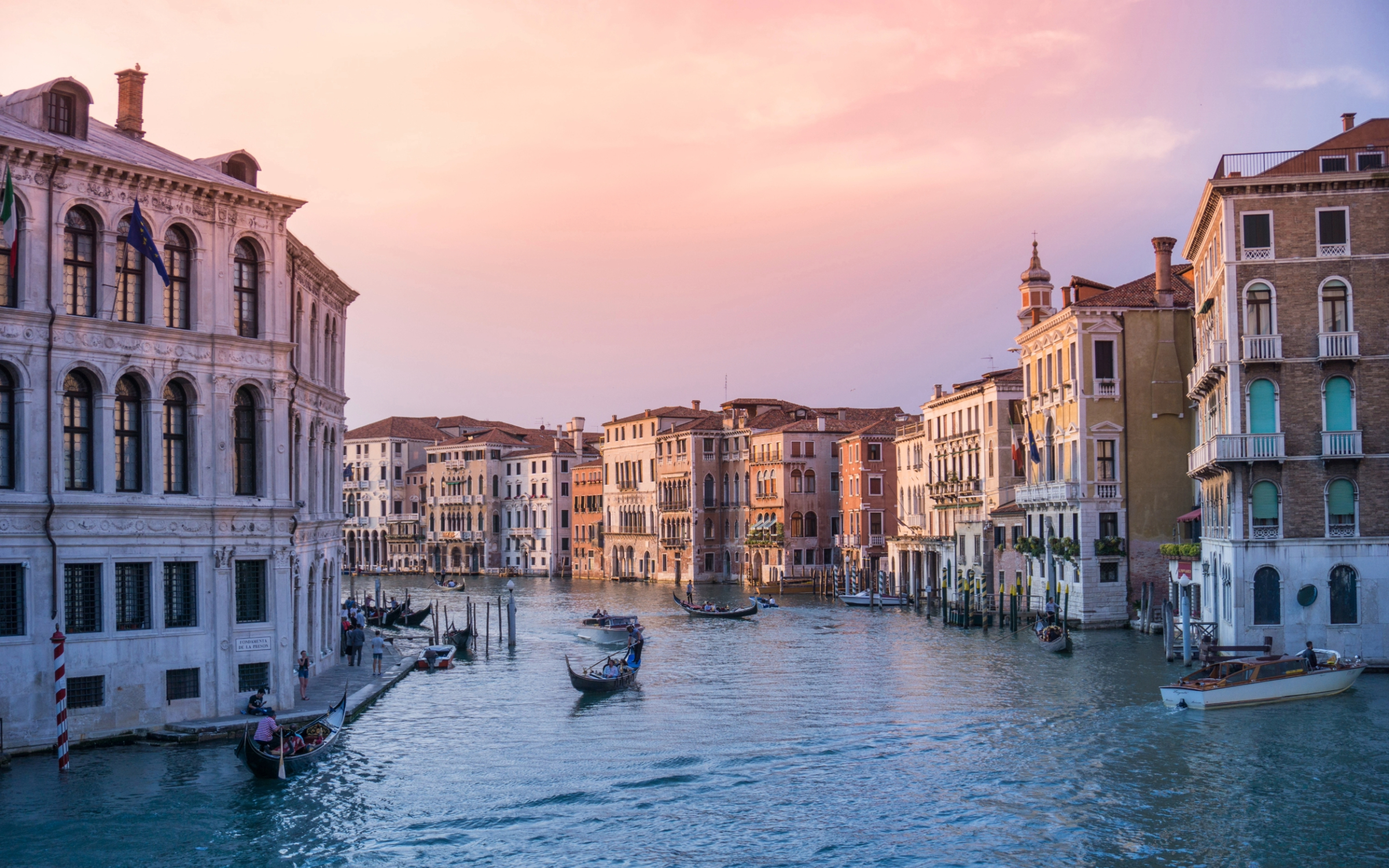 Several gondolas on a wide canal in Venice surrounded by old buildings under a pink and orange sky.