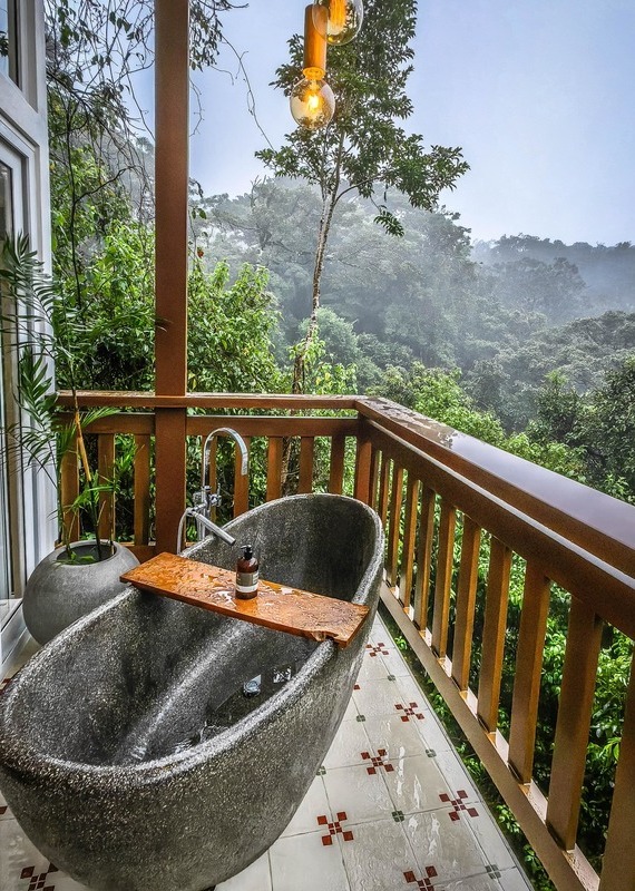 A grey stone soaking tub on a tiled balcony with a wooden railing overlooking a foggy rainforest.