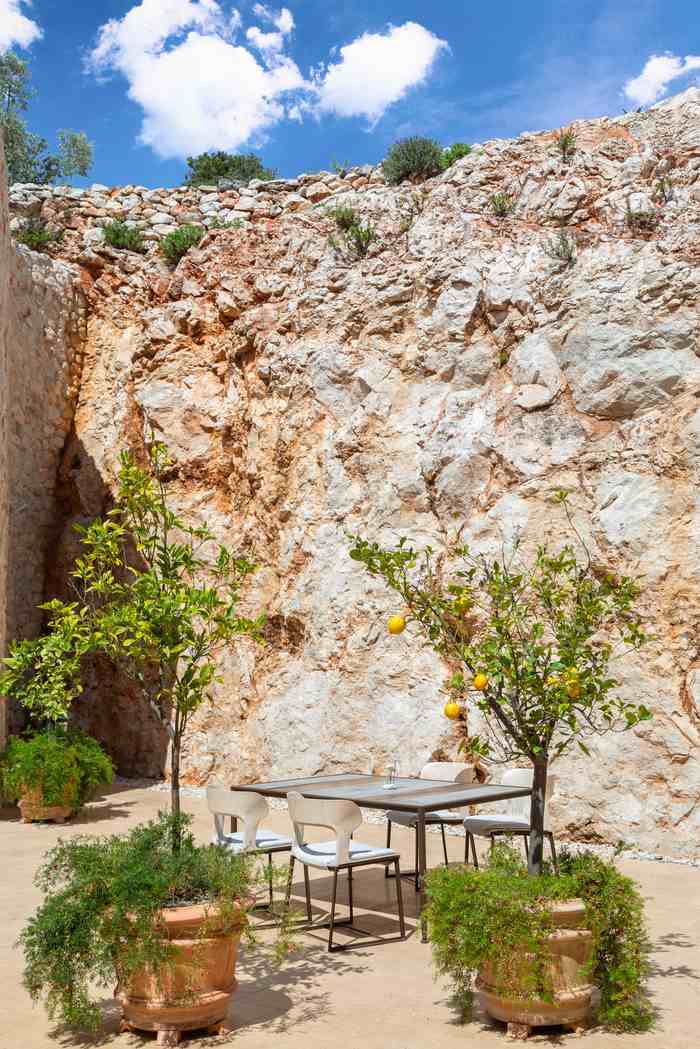A modern dining table and chairs on a stone terrace in front of a tall, dramatic rock wall.