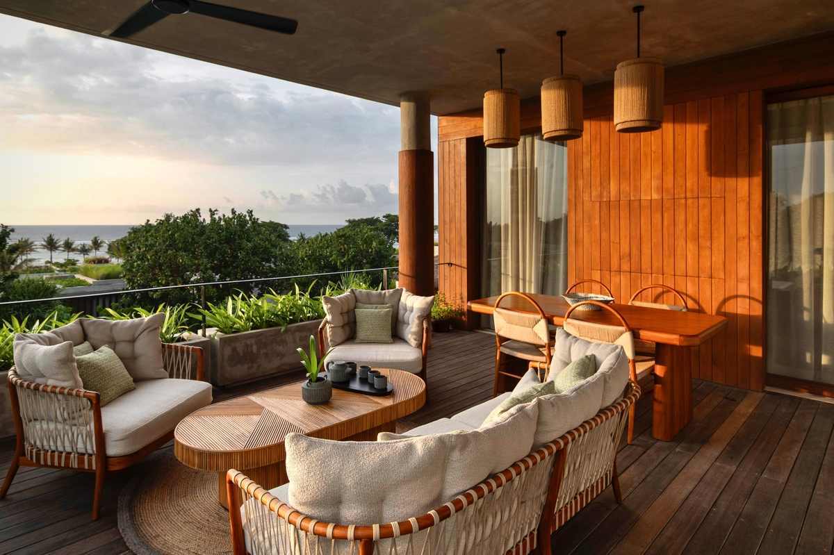 A covered wooden balcony with a dining table, comfortable armchairs, and views of the ocean at dusk.