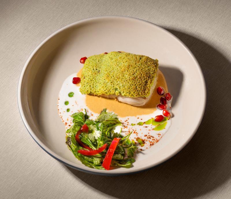 A high-angle shot of a plated fish dish with green crust, orange sauce, pomegranate, and greens.
