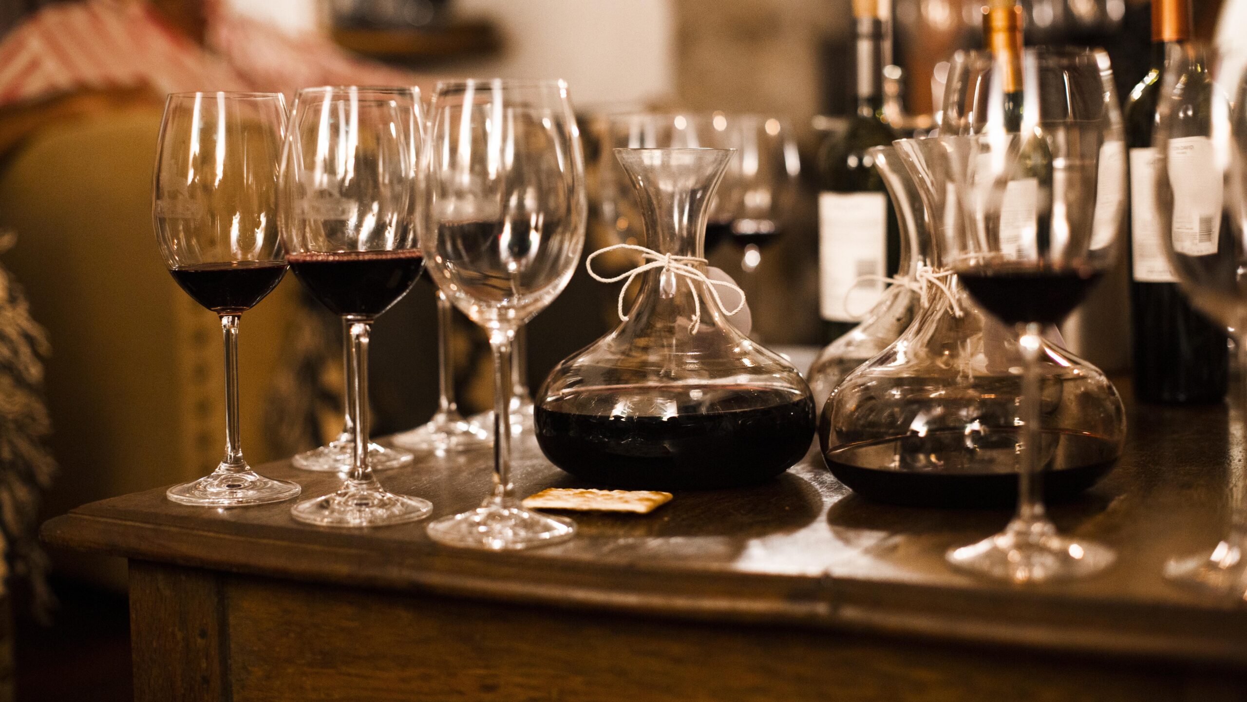 Red wine in glasses and decanters on a wooden table, with bottles in the background.