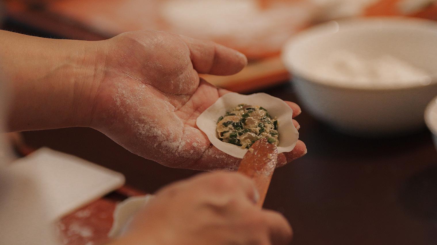 Hands preparing a dumpling with filling, dusted with flour over a wooden table.