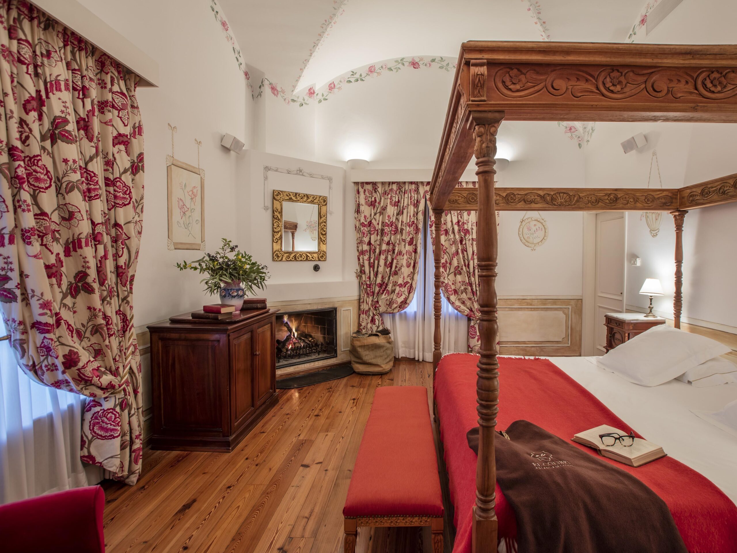 Elegant bedroom with four-poster bed, red accents, fireplace, and floral drapes.
