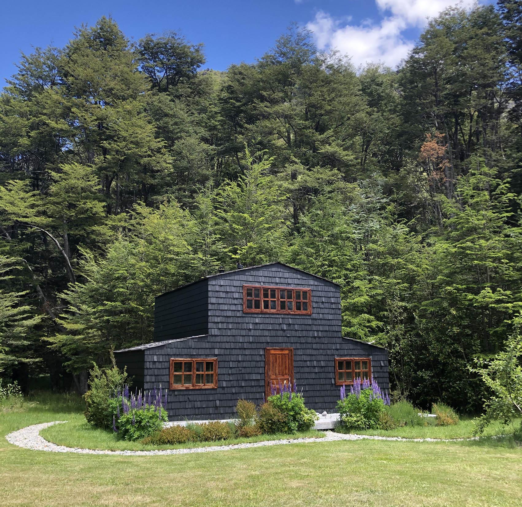 A two-story dark wooden cabin with a manicured lawn and forest backdrop under a blue sky.