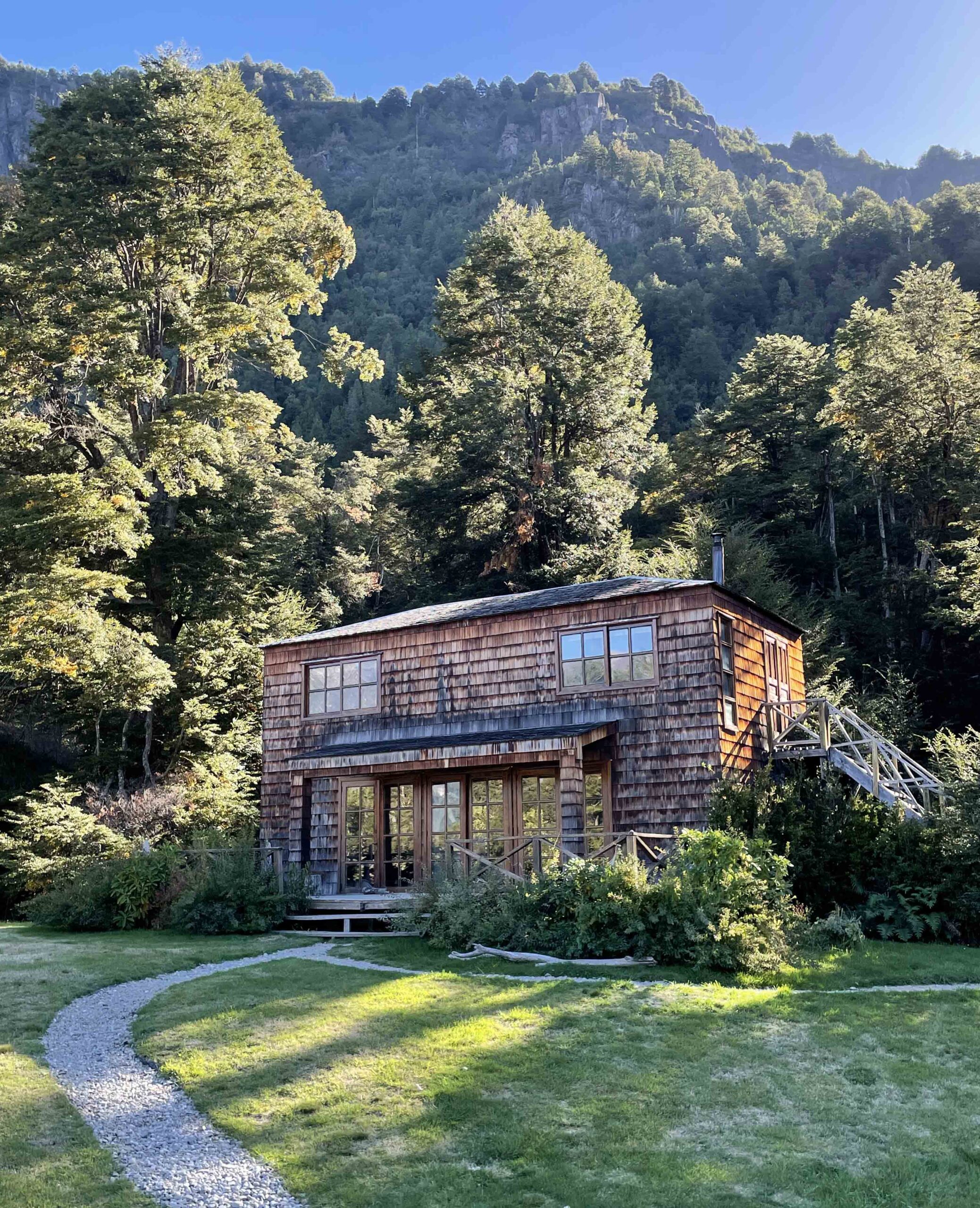 A wooden cabin surrounded by forest with a mountain backdrop under a clear sky.