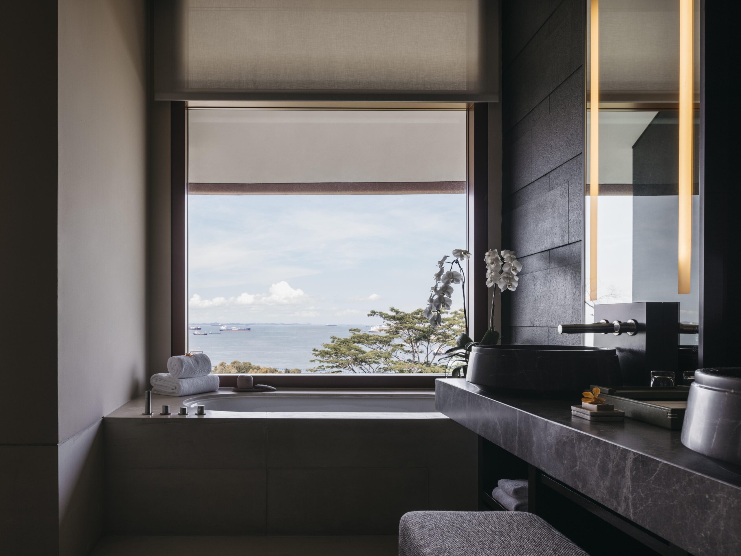 Modern bathroom with large window overlooking the ocean, featuring dark stone finishes and orchids.