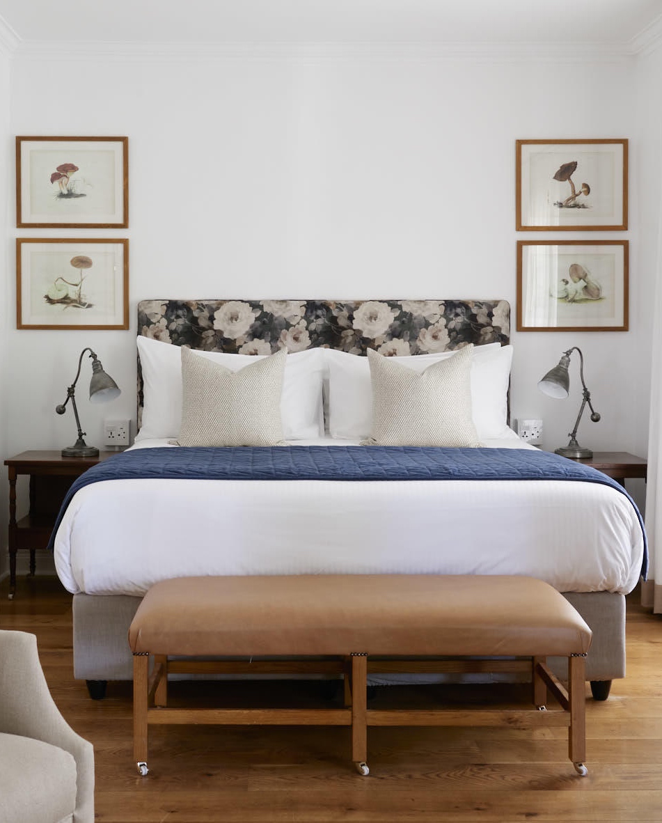 Elegant bedroom with floral headboard, white bedding, navy throw, artwork, and bedside lamps.