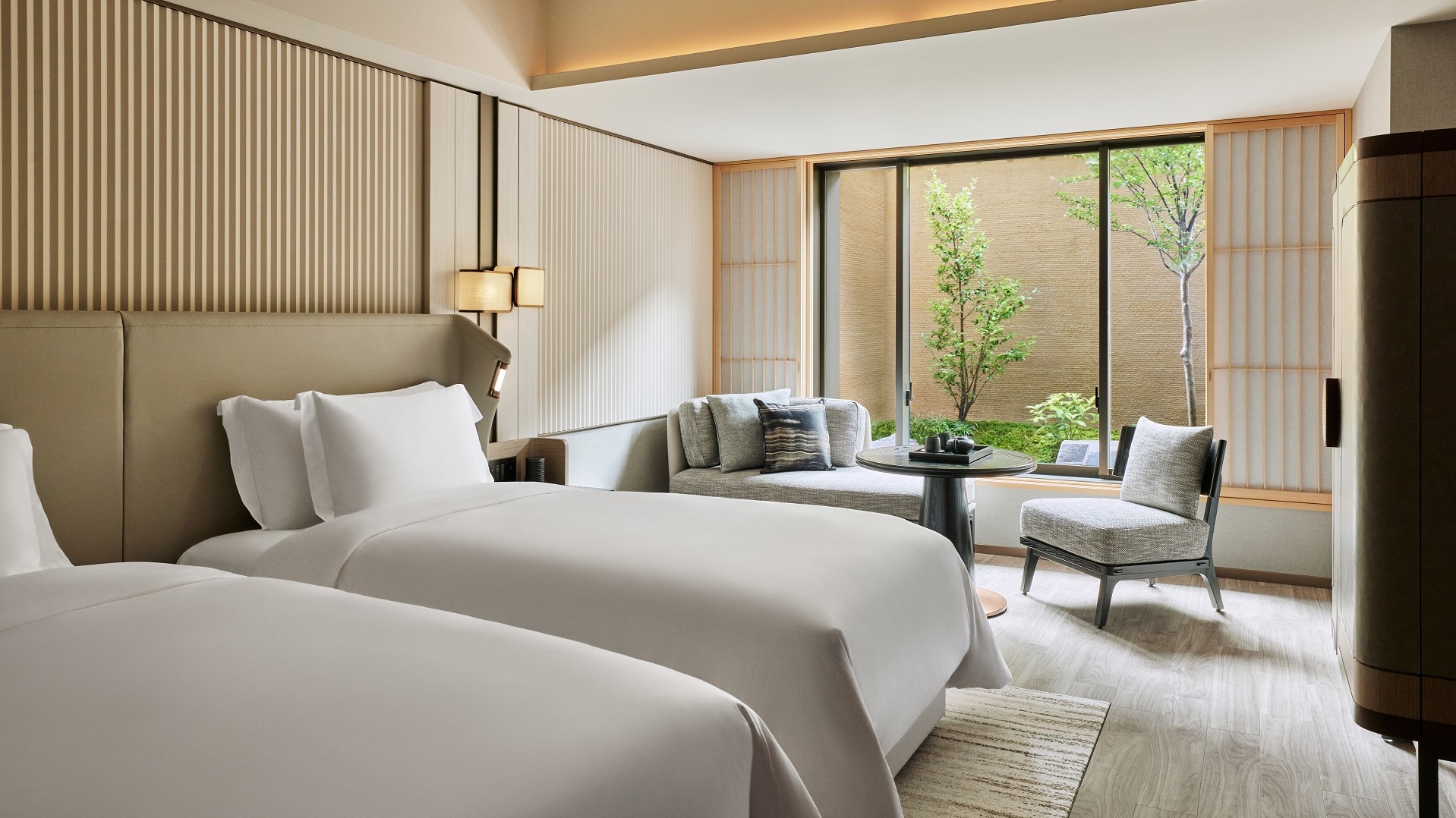 Modern hotel room with twin beds, minimalist decor, and a view of a small garden.
