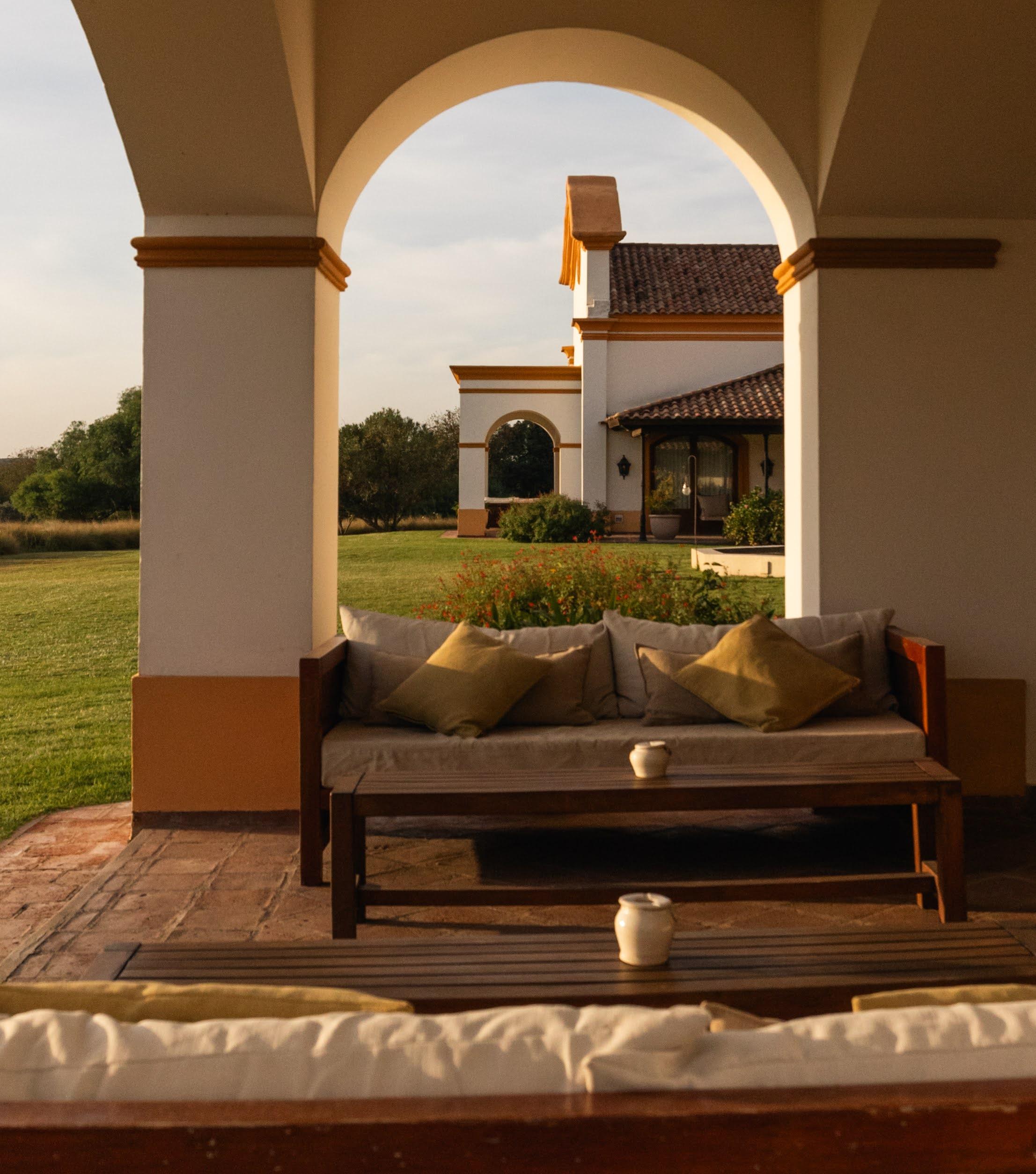 A cozy outdoor seating area with a wooden sofa and cushions, framed by arches overlooking a garden.