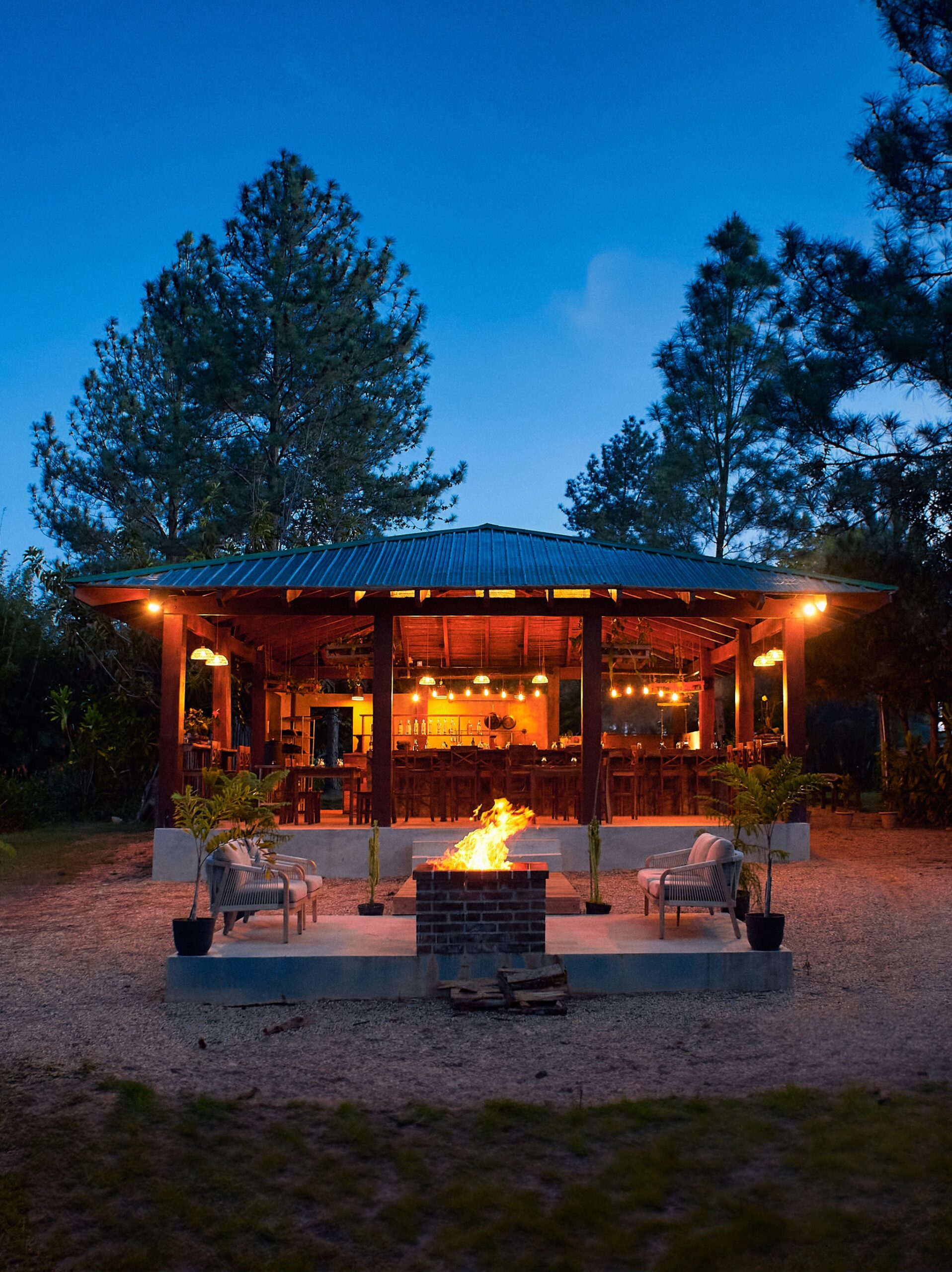Outdoor patio with lit fire pit at dusk, surrounded by trees and a cozy seating area.