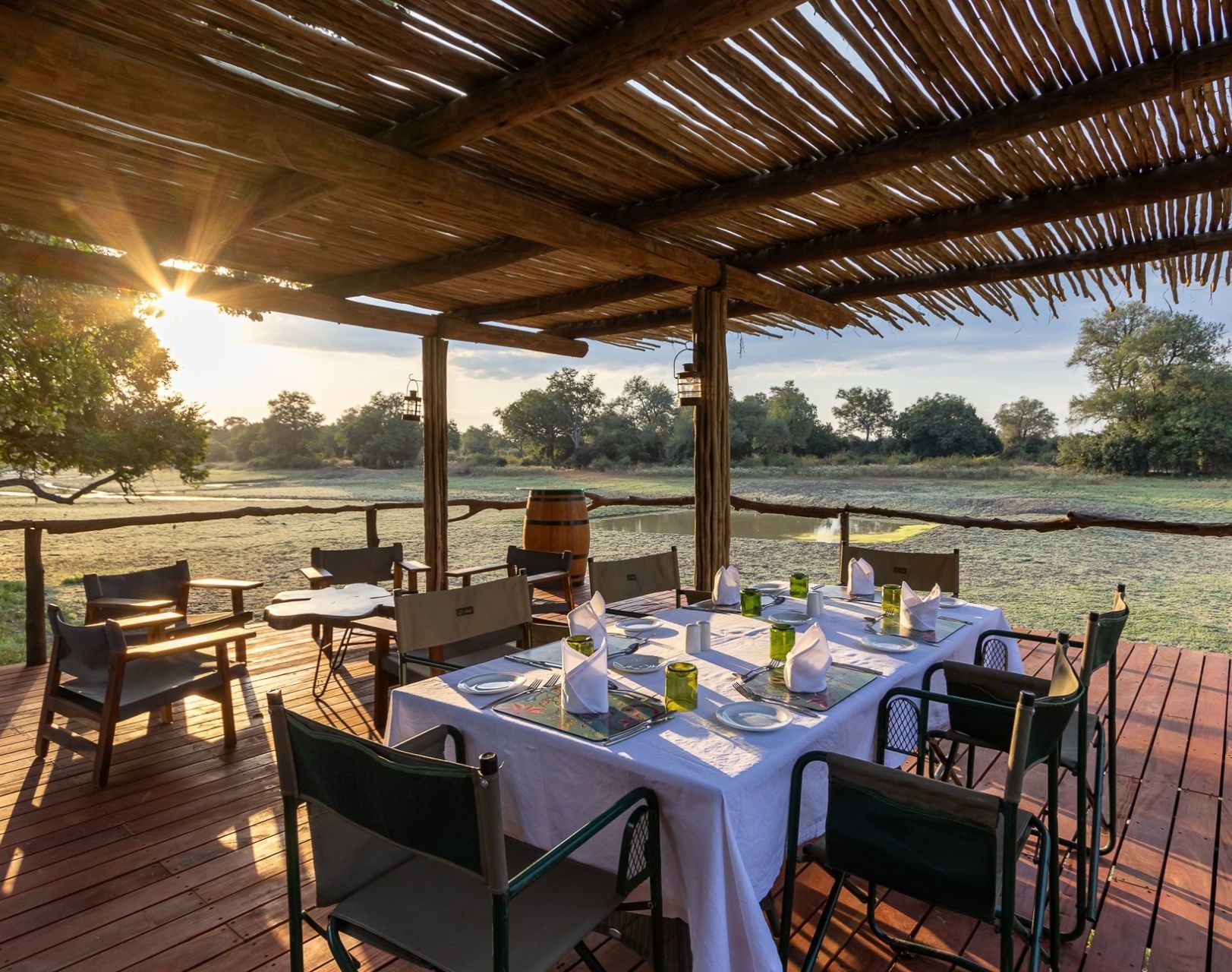 Outdoor dining setup on a wooden deck with a thatched roof at sunset, overlooking a scenic savanna.