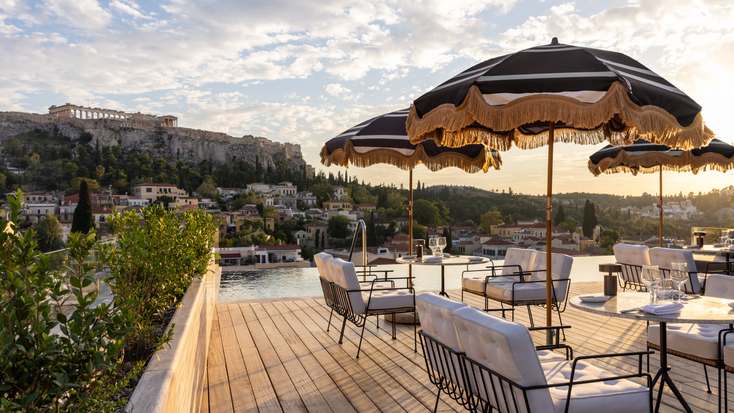 Elegant rooftop seating with striped umbrellas overlooks Athens, featuring rich greenery and the Acropolis in the background during sunset.