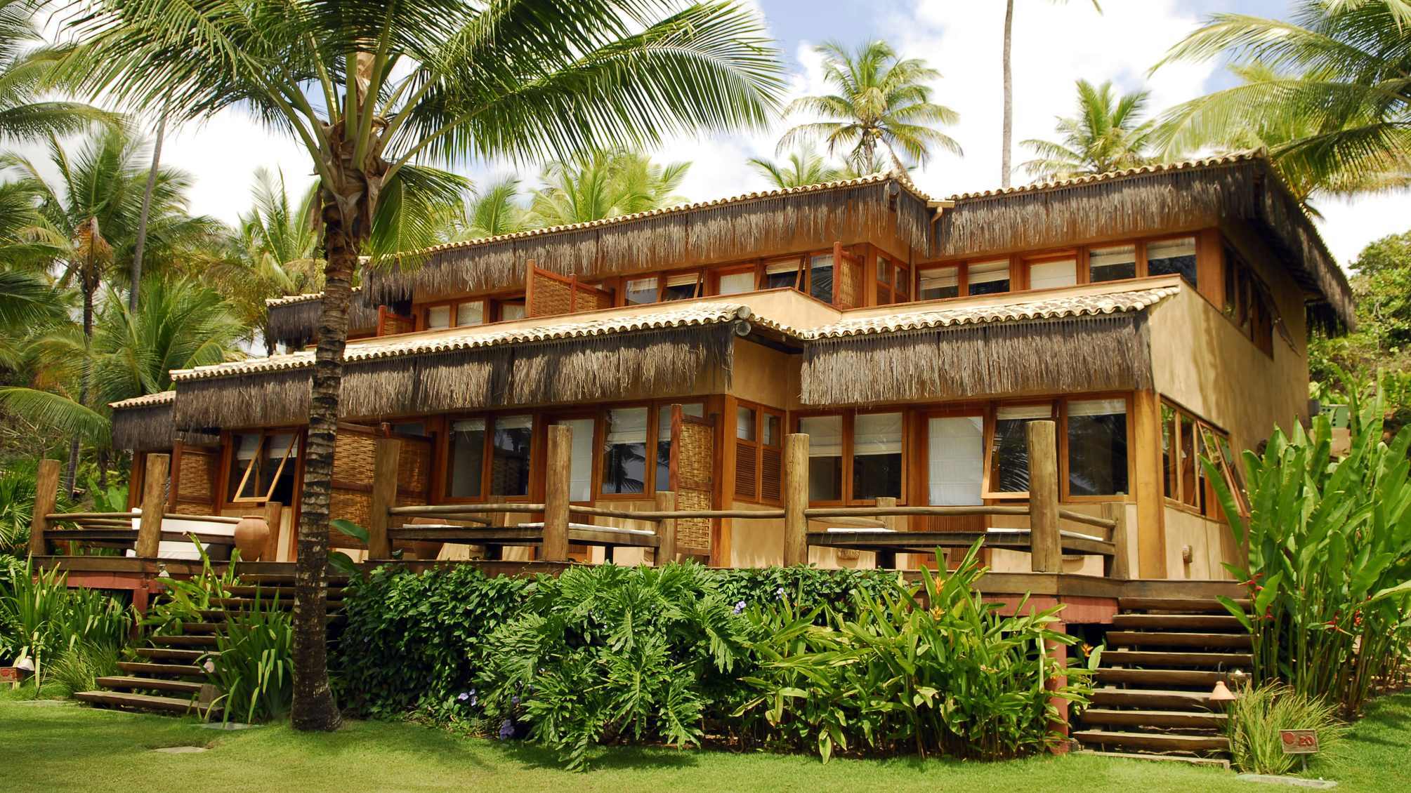 Tropical wooden house with thatched roof, surrounded by palm trees and lush greenery, featuring balconies and wooden stairs.