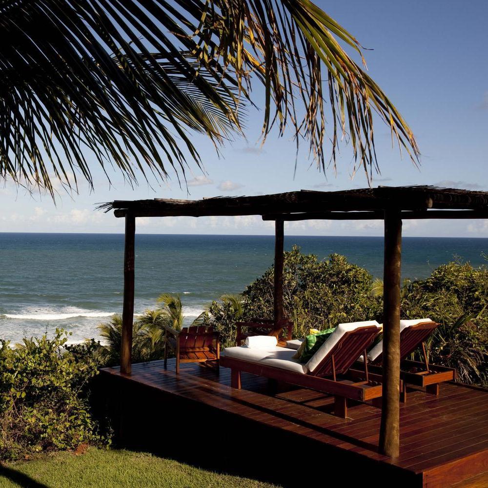 A serene ocean view from a wooden deck featuring two lounge chairs and lush tropical greenery under a clear blue sky.