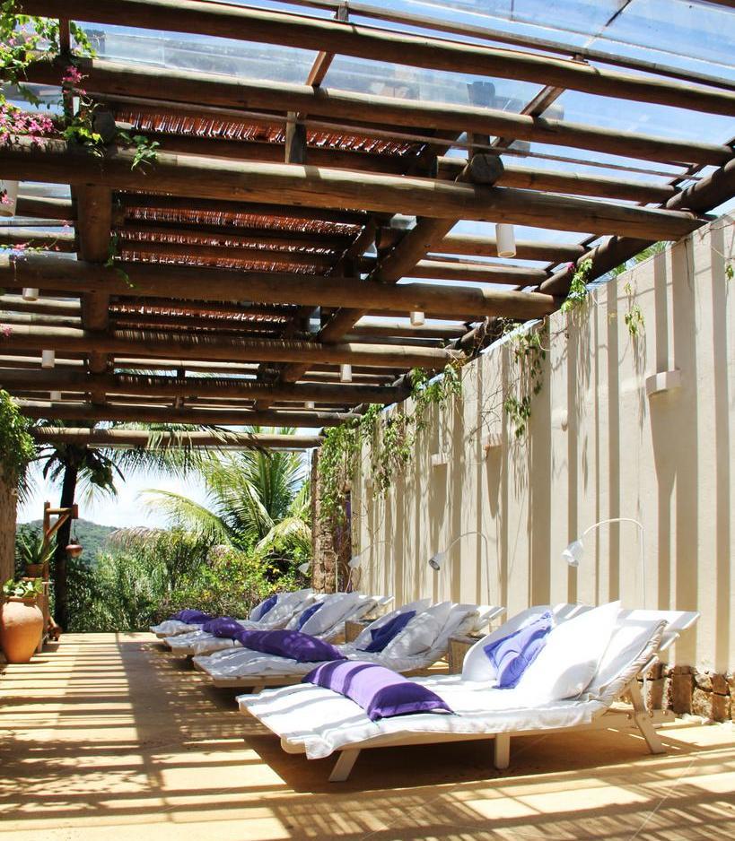 Outdoor lounge area with white sunbeds adorned with purple pillows, surrounded by greenery and a wooden canopy letting in sunlight.