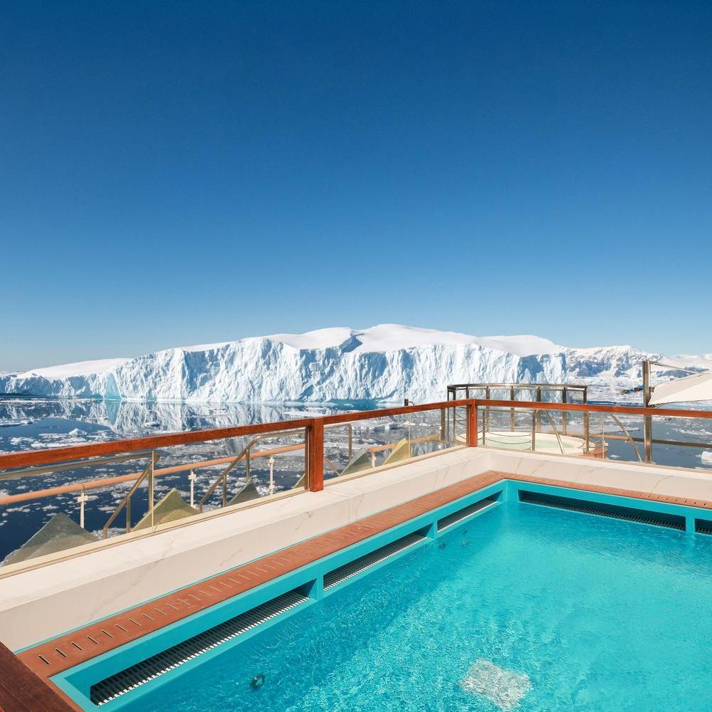 Bright blue rectangular swimming pool on a ship deck, overlooking a massive tabular iceberg floating in calm icy waters.