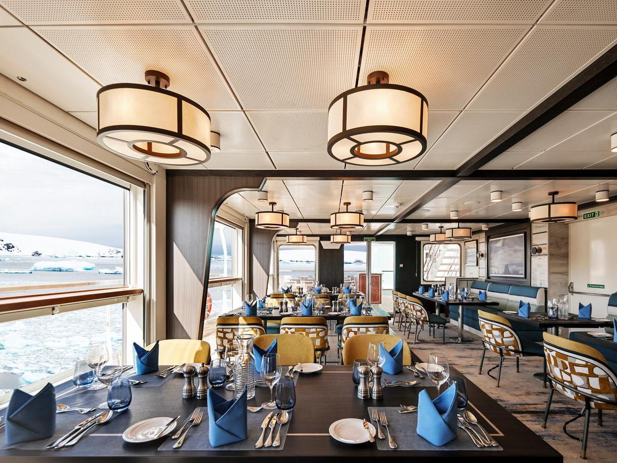 Bright dining room with set tables, blue napkins, and large windows looking out at an icy seascape.