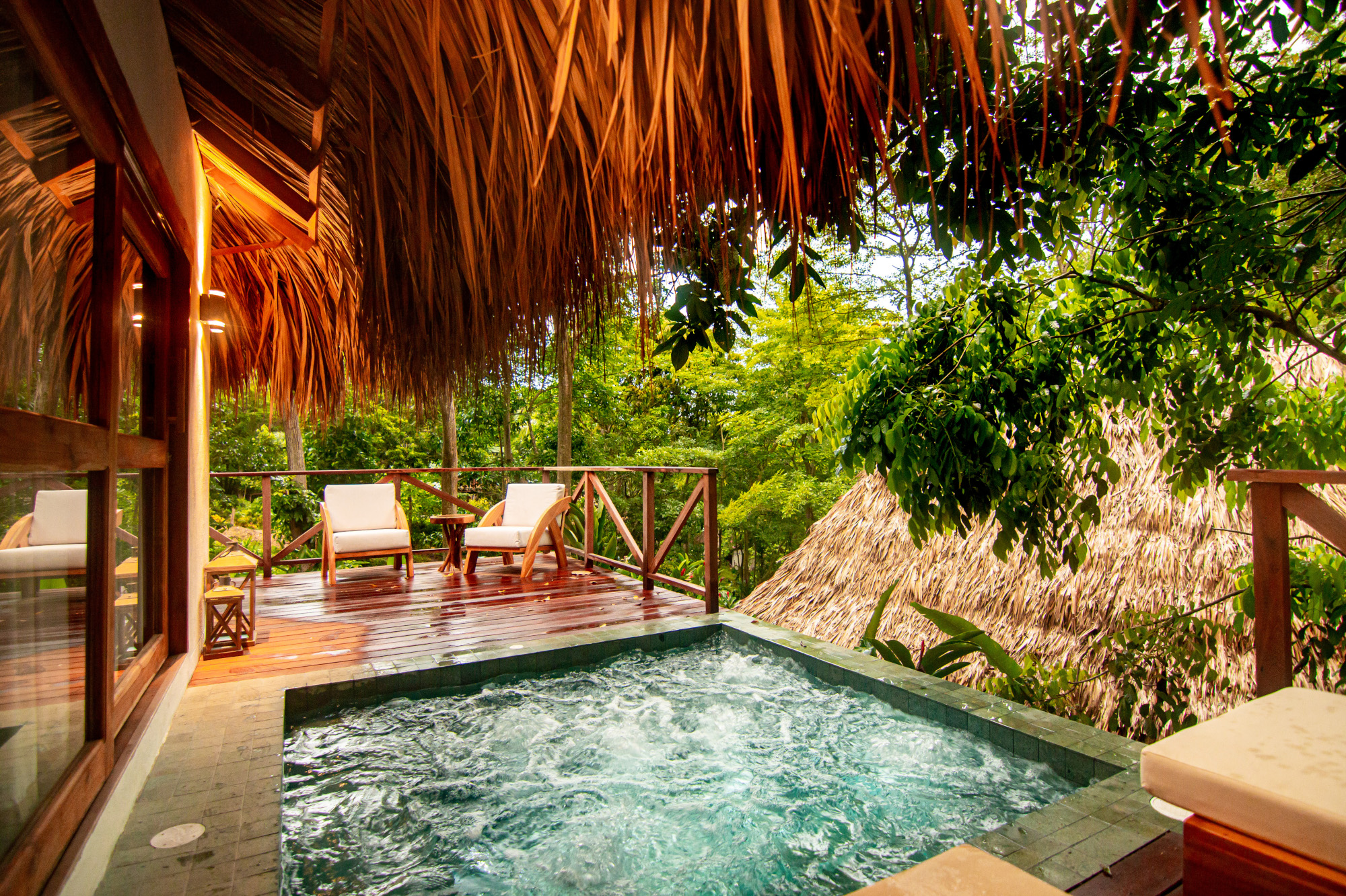 In the foreground, a small pool with green water and overhanging thatched roof. Around it is a wooden deck with two white lounge chairs, and in the background is dense green jungle.