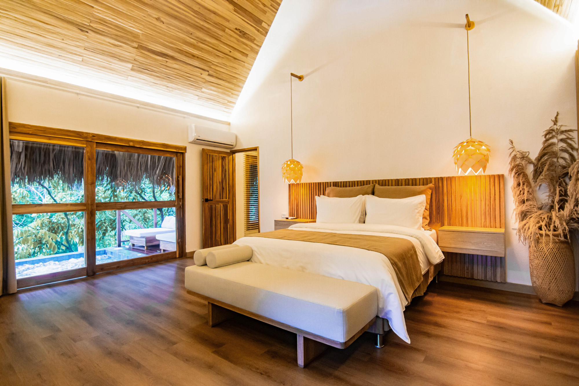 A bedroom with a double bed with white and brown sheets and pillows and a hardwood headboard and flooring. The room has white walls and a hardwood ceiling, and there is a large picture window on the left looking out into the jungle.