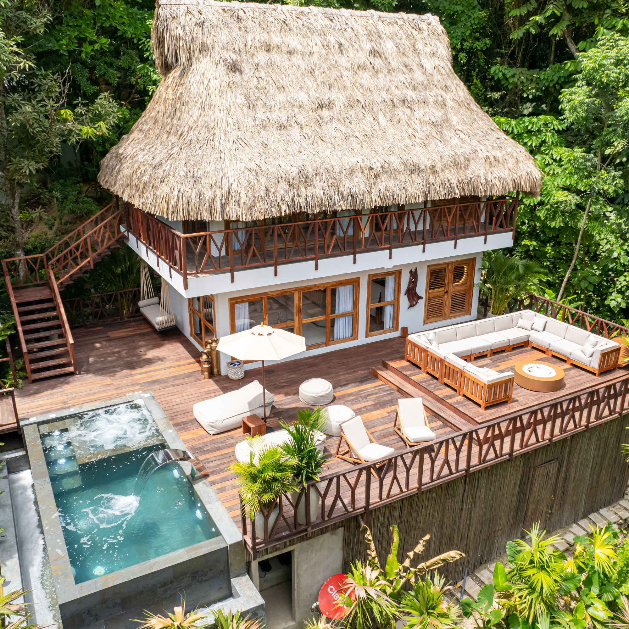 A view from above of a two-storey room with whitewashed walls, wooden railings and stairs and a thatched roof. Around it in the foreground is a deck with white sofas, lounge chairs and a small pool with blue water. In the background is dense green foliage.