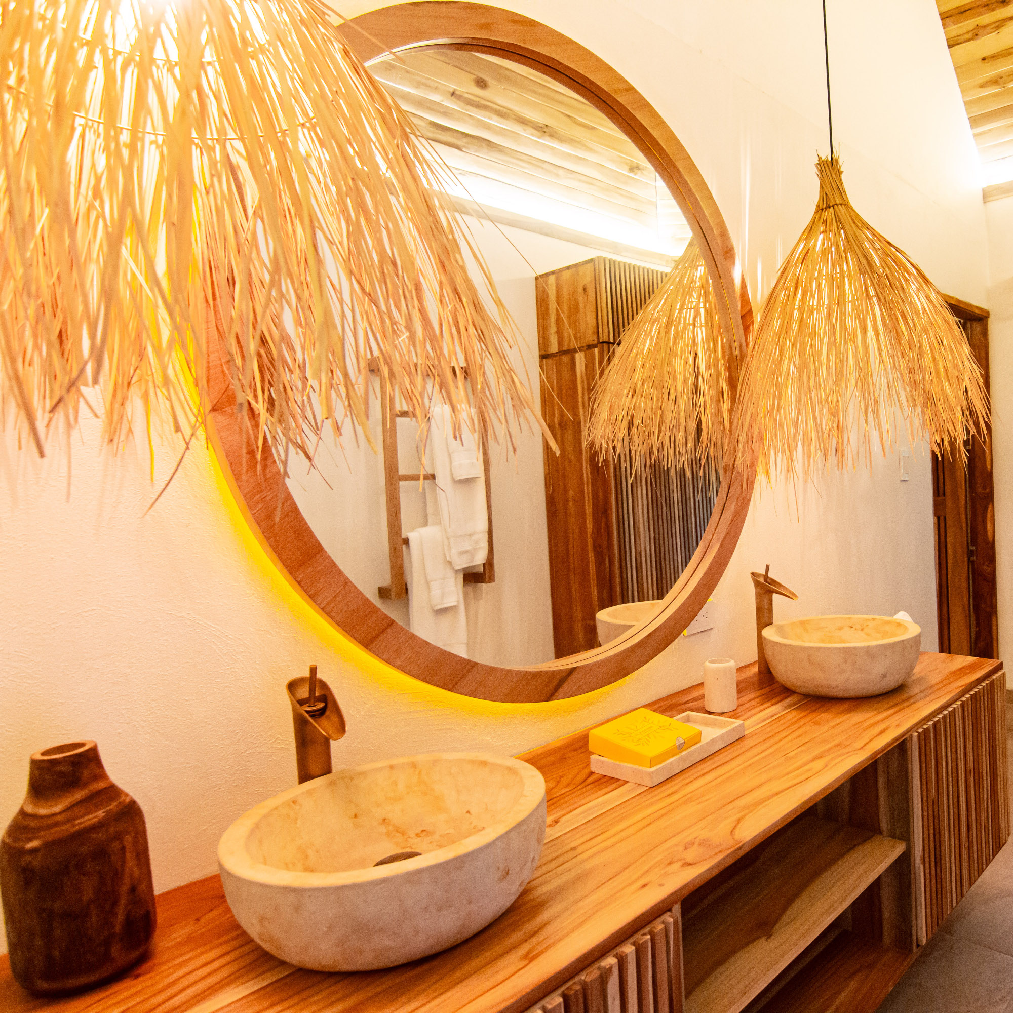 A bathroom vanity with a big round mirror, two marble sinks set on a wooden countertop with a dark bottle on the left. Overhead are two lamp shades made of brown grass.