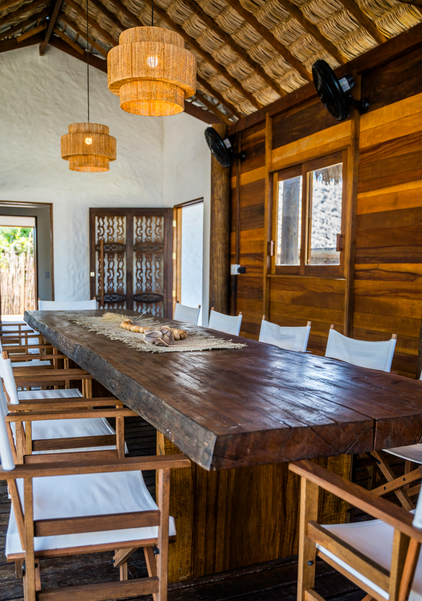 A long dark wooden dining table with white chairs sits in a room with one wooden wall and one white wall, two basket lamps hanging overhead and an open doorway at the far end.