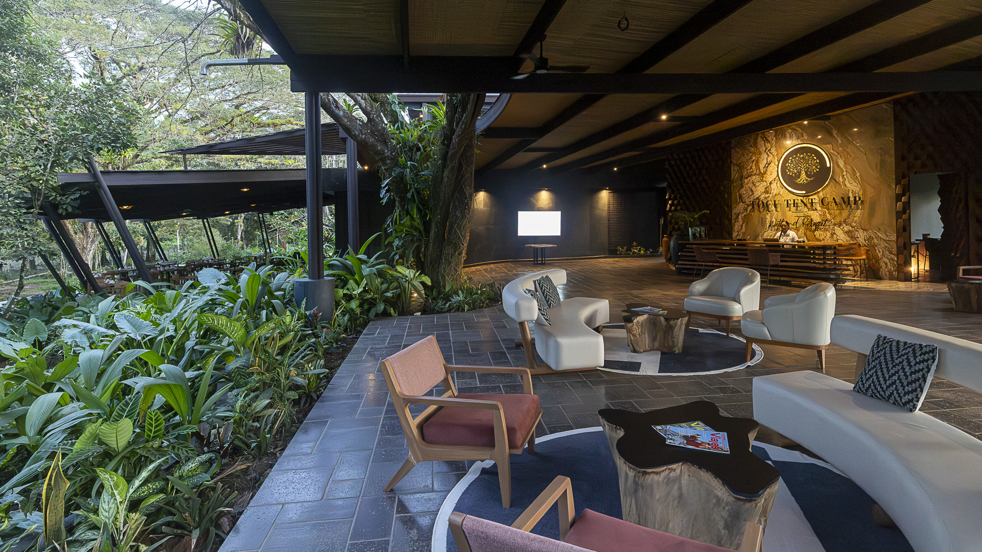 An open-air hotel reception area with a covered wooden deck where there are lounge chairs and sofas overlooking jungle greenery.