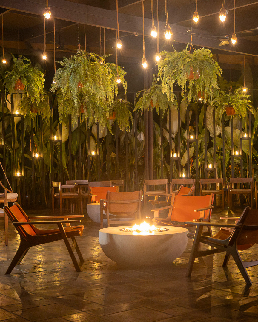 A bar scene with tables and orange chairs facing a lit firepit. Overhead are baskets of hanging green ferns and bare lightbulbs.