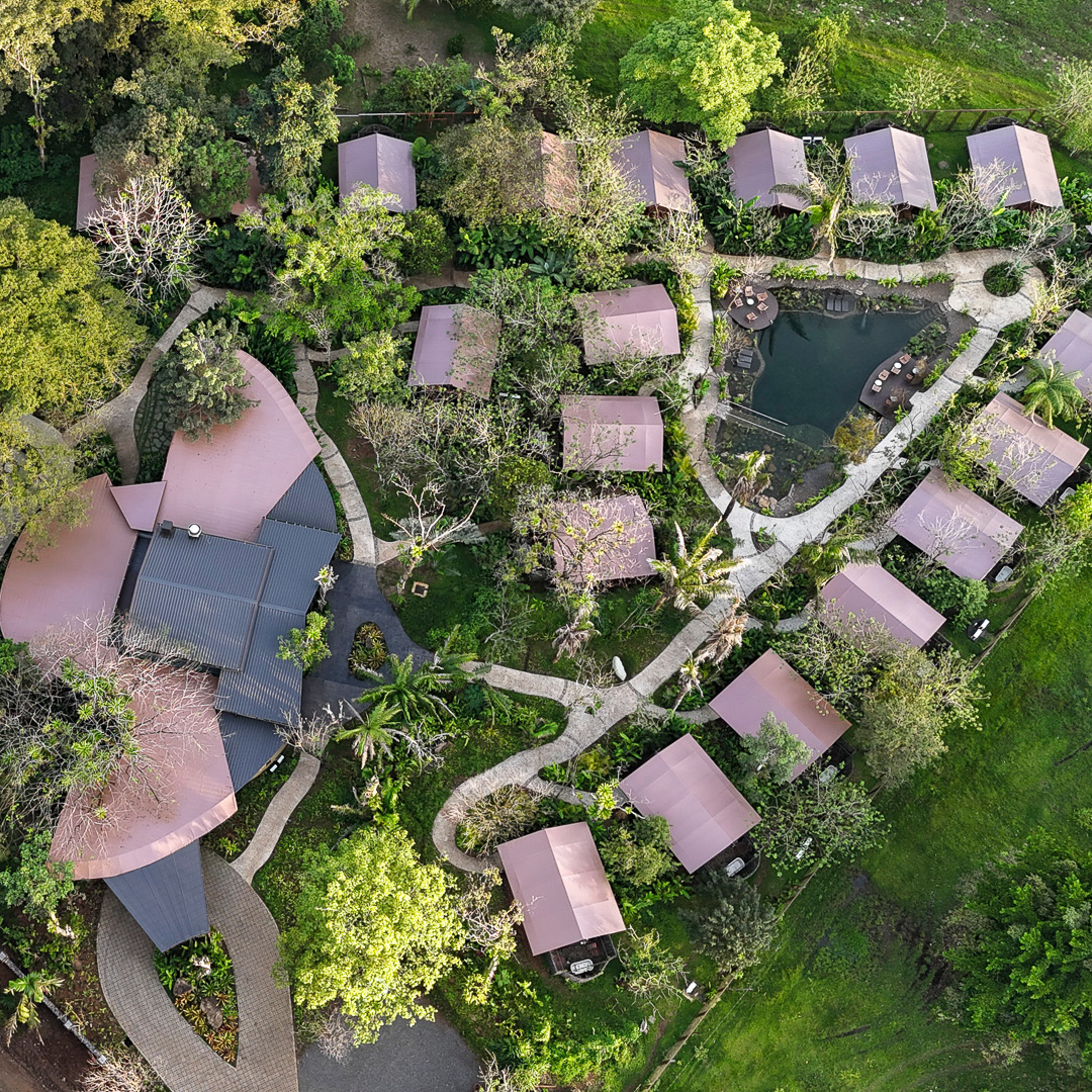 An aerial shot of a hotel with a main lodge building on the left and 18 smaller buildings can be seen to the right with some small walkways connecting them and lots of green trees and grass in between them.