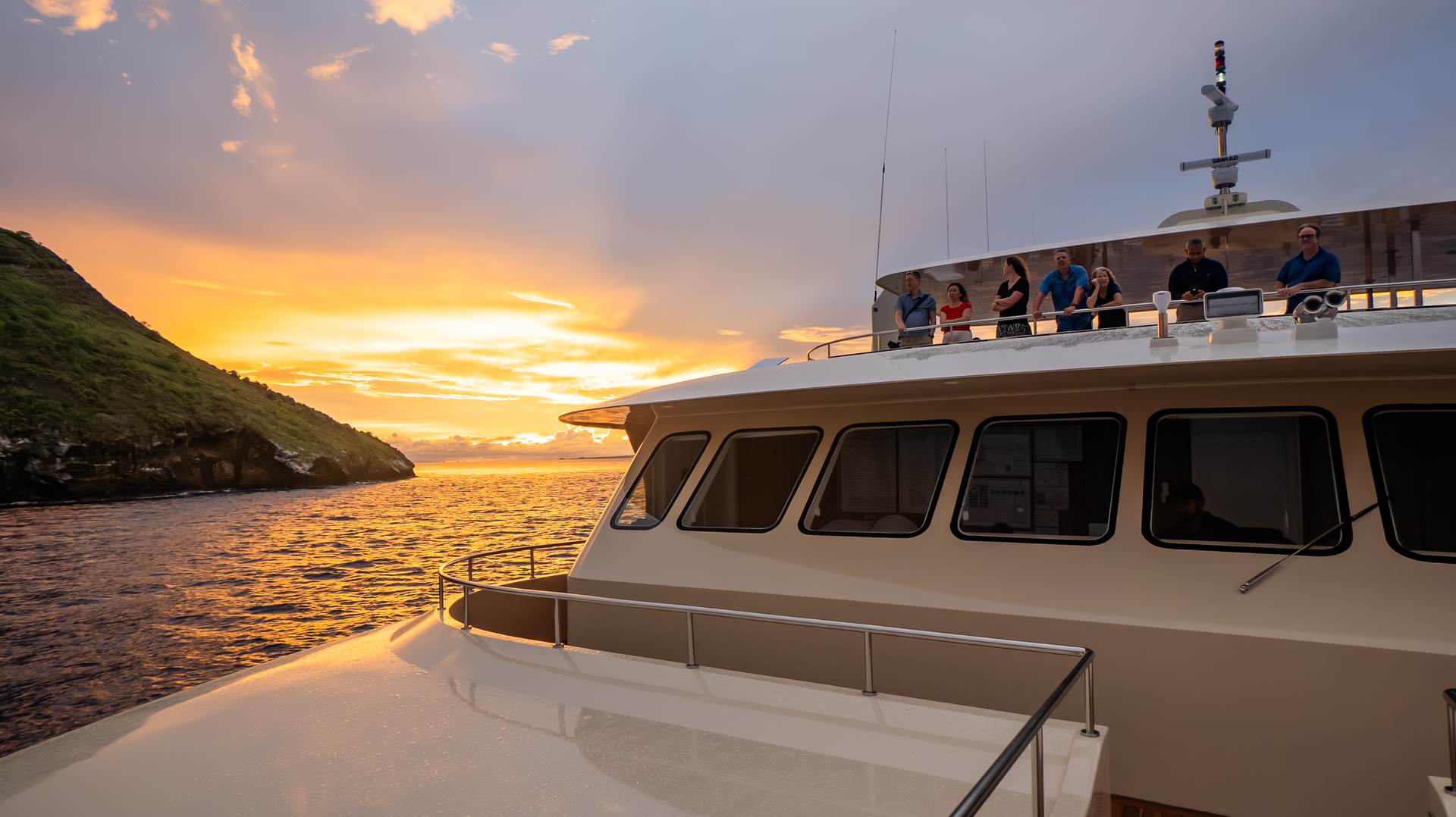 Passengers stand on a yacht upper deck looking out at a vibrant orange and yellow sunset over the water.